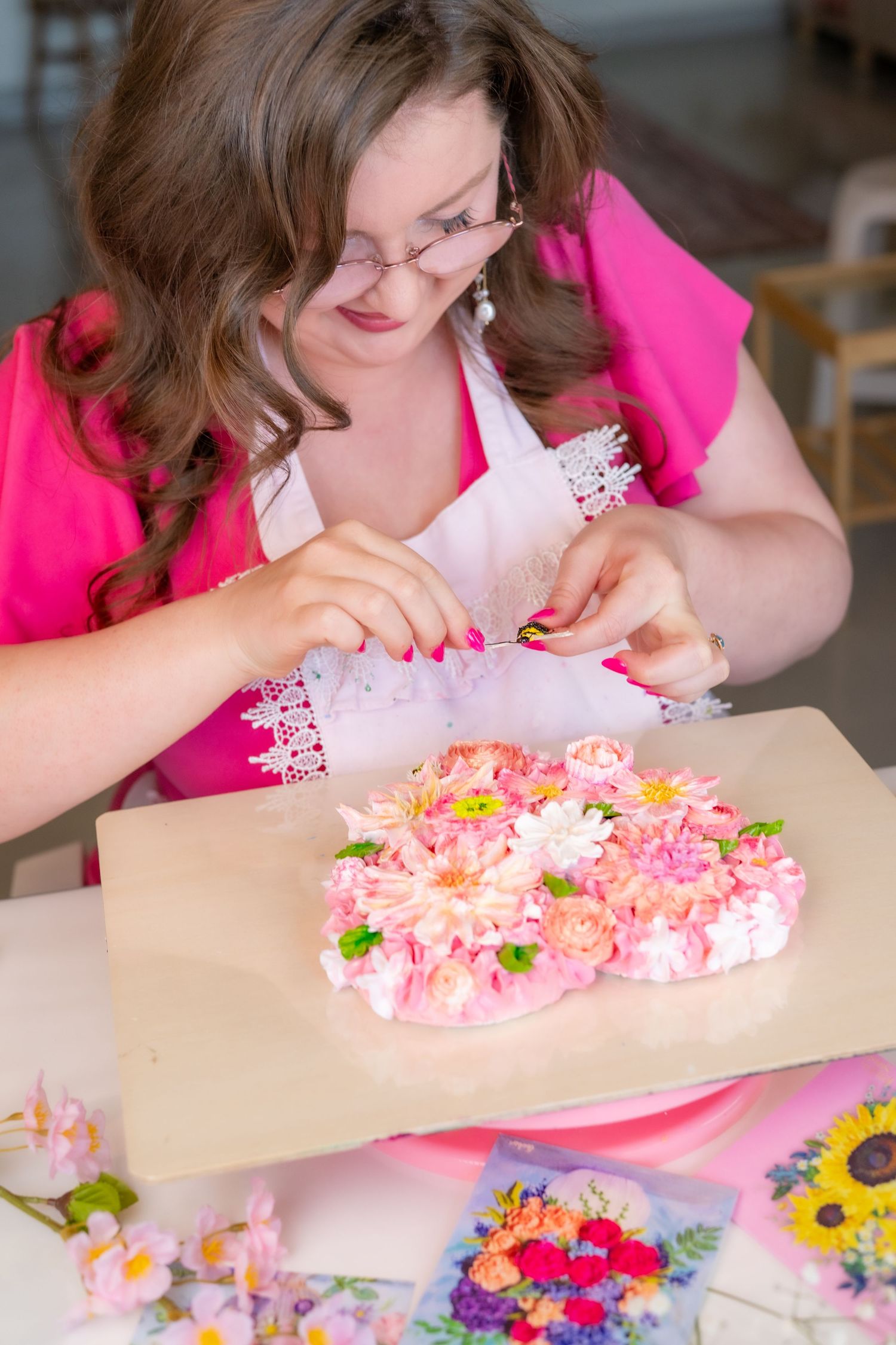 Woman in pink decorating a floral cake with fresh flowers at a table covered in blooms.