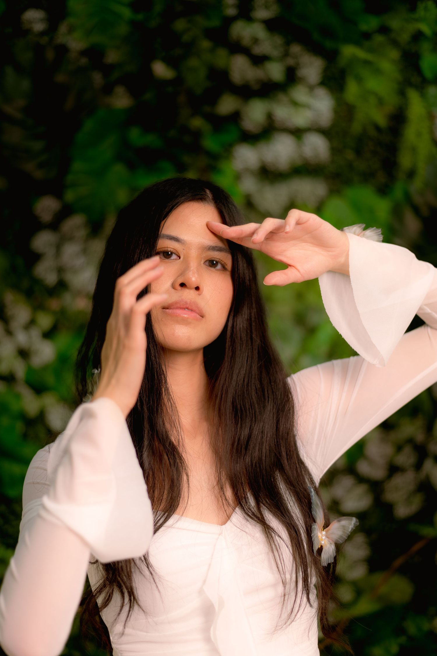 Woman in white long sleeve top poses against lush green garden backdrop.