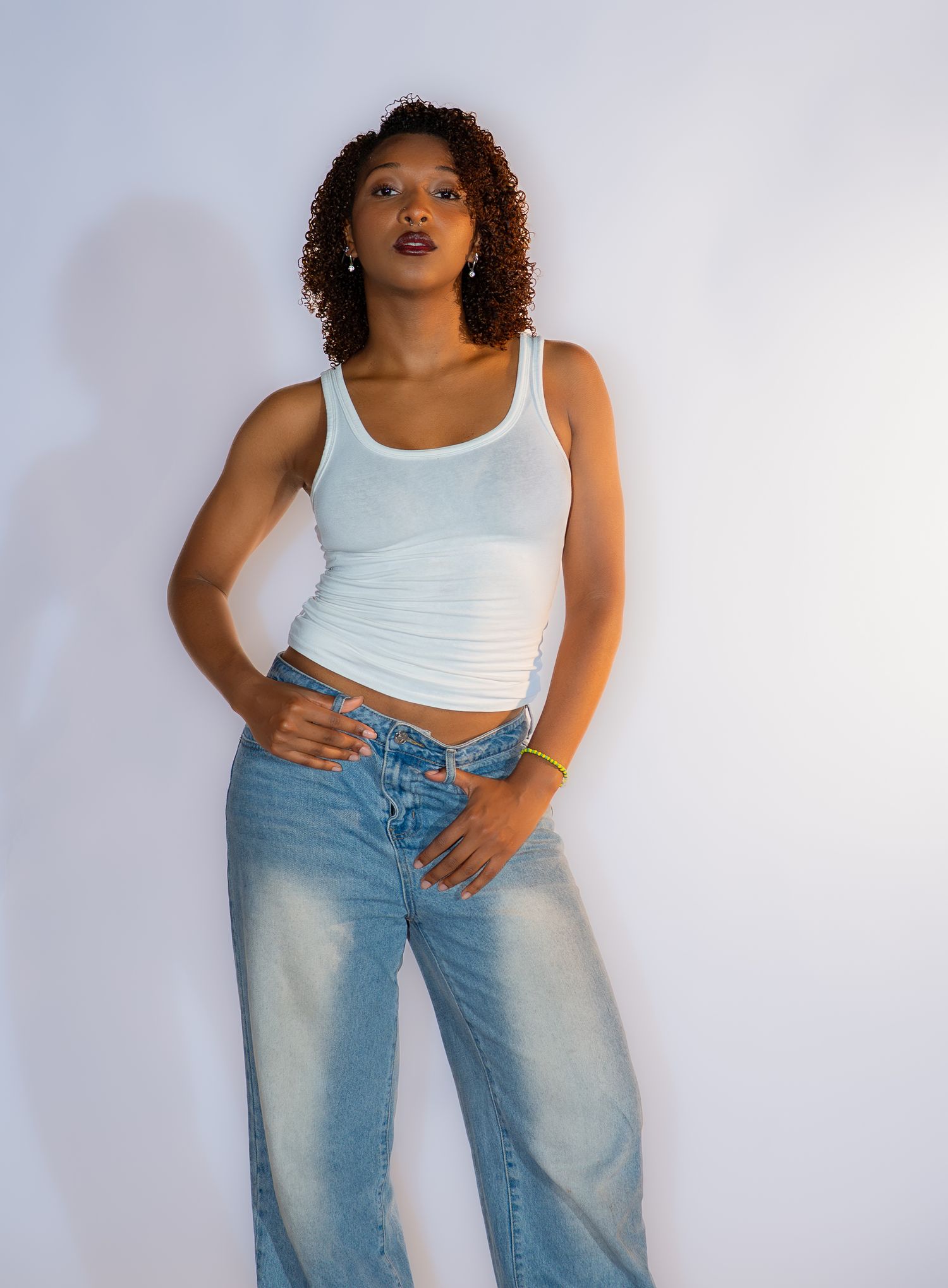 A person in a white tank top and light wash baggy jeans poses against a white background in a fashion photoshoot.