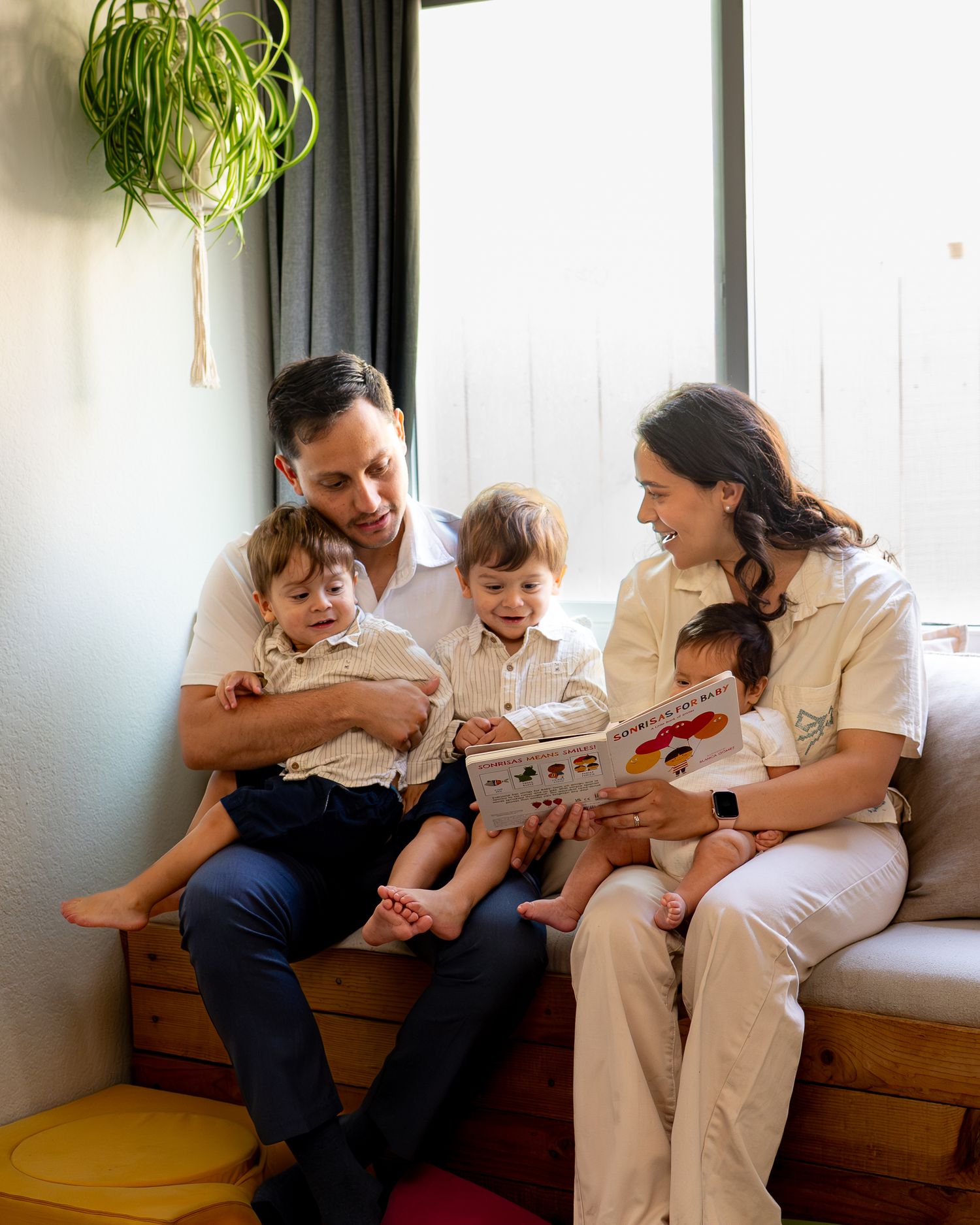 A happy family of five sits together on a bench, reading a colorful book, dressed in matching cream outfits.