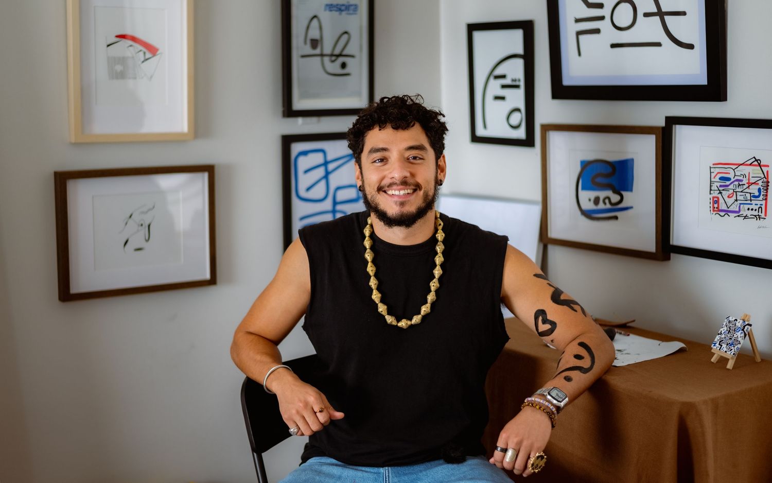 A person wearing a black sleeveless shirt sits at a desk with framed artwork displayed on the wall behind them.