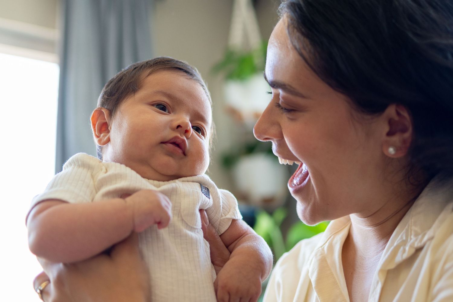 A smiling mother holds her newborn baby up close, sharing a tender moment in a bright, cozy home setting.