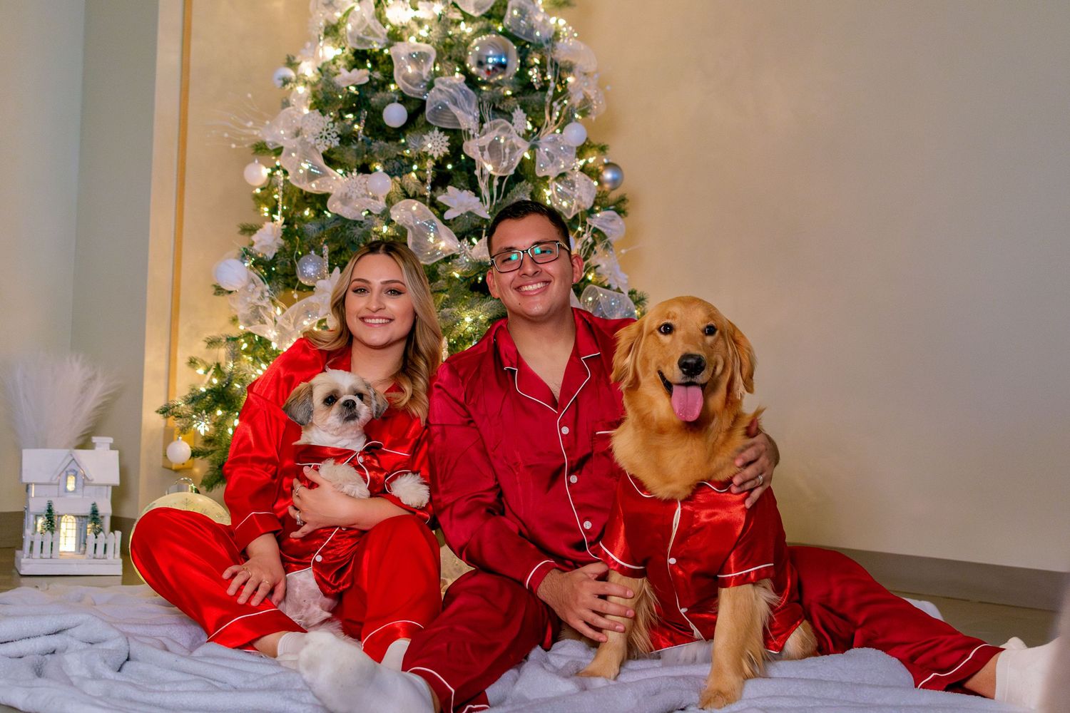 Couple in matching red Christmas pajamas sitting with a Chihuahua and Golden Retriever in front of a decorated tree.