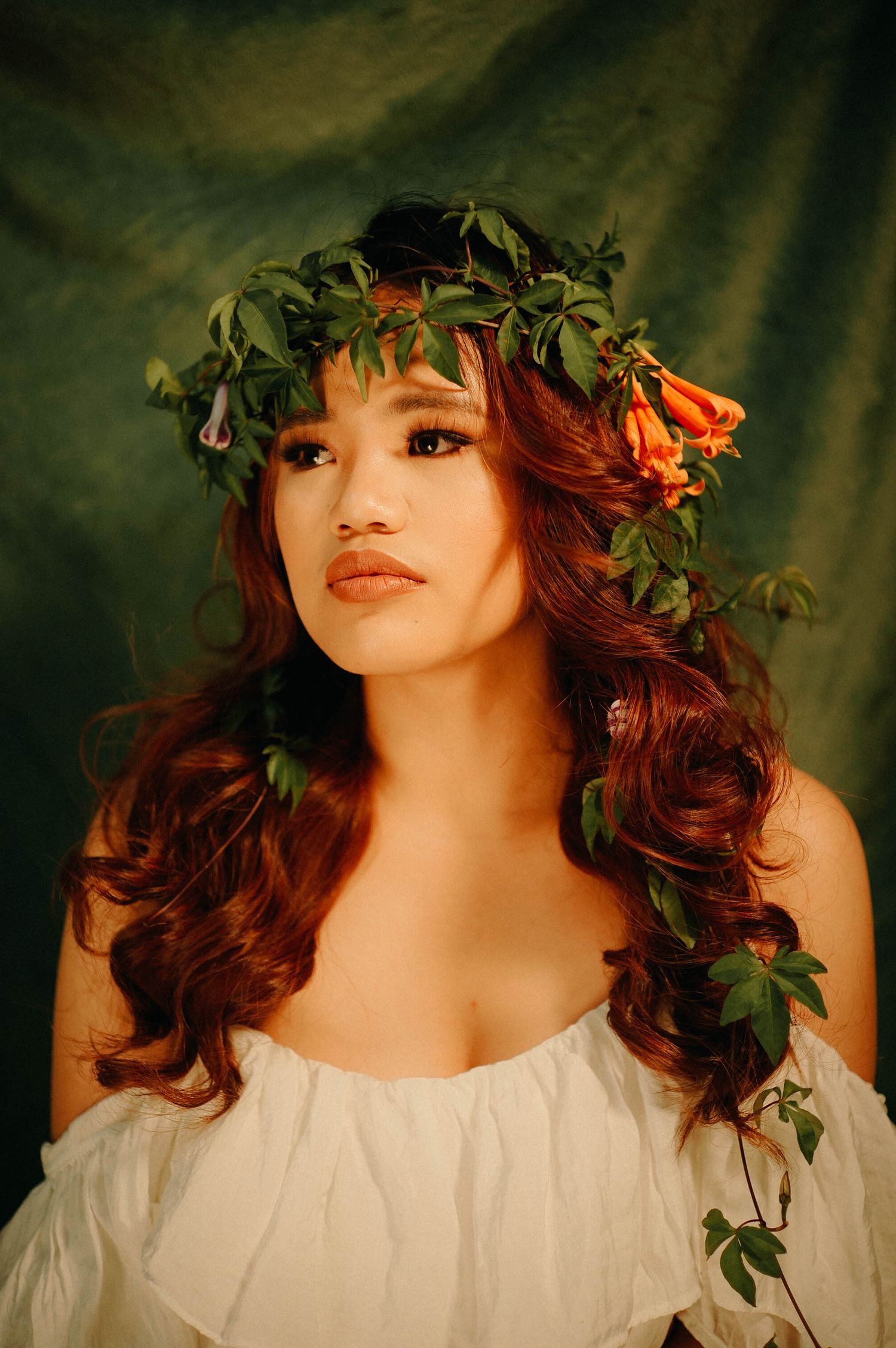 Ethereal portrait with flower crown and flowing red hair against dark green background.