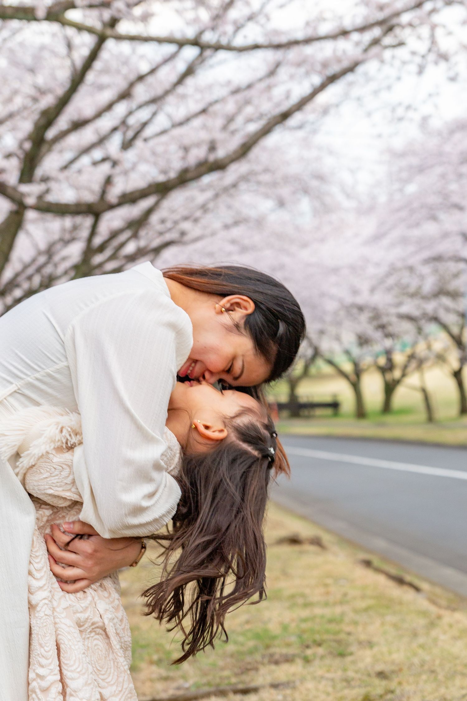 A person in a white sweater dances joyfully under blooming cherry blossom trees along a rural road.