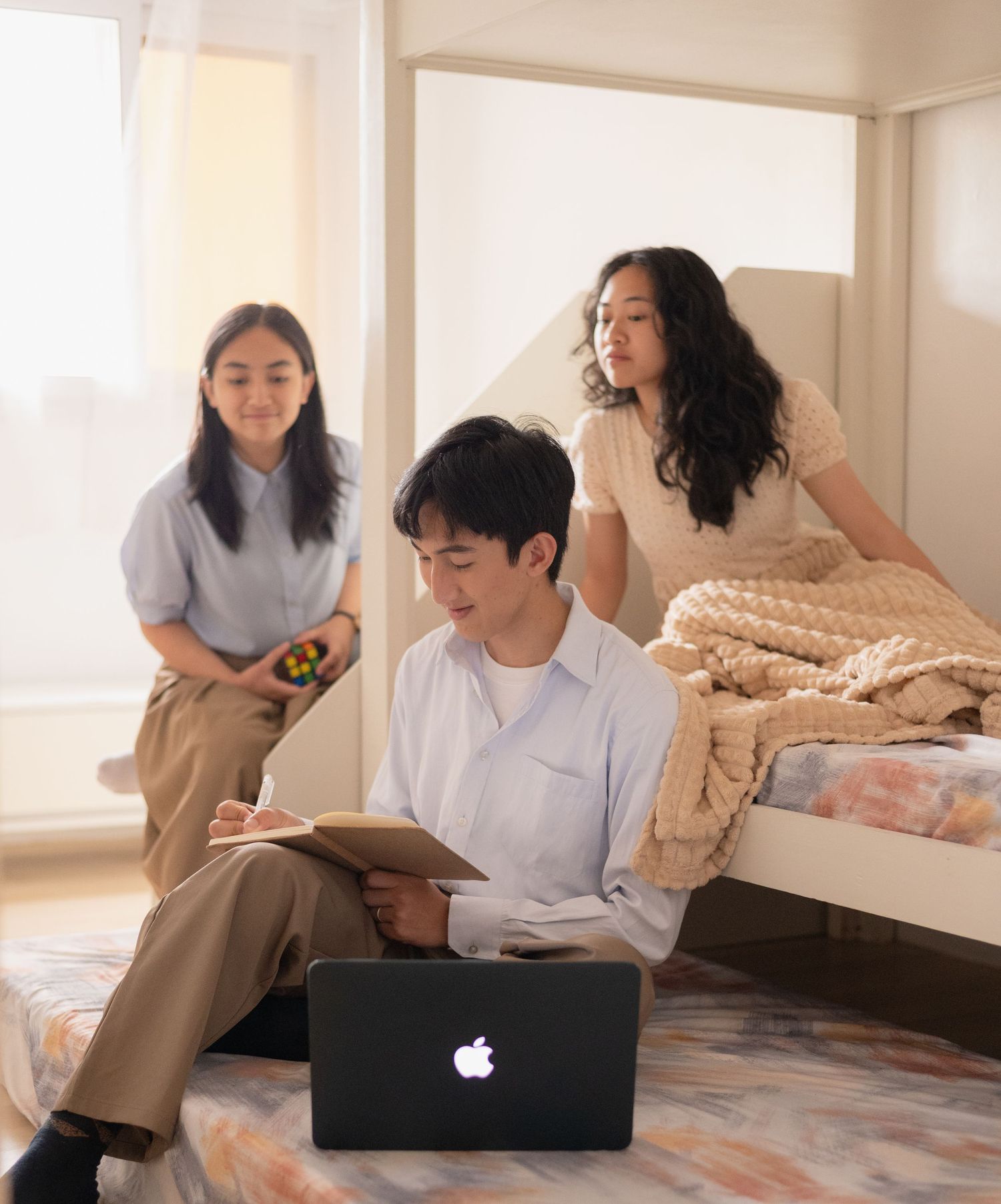 Three young adults relaxing in a sunlit bedroom with a MacBook, book, and Rubiks cube.