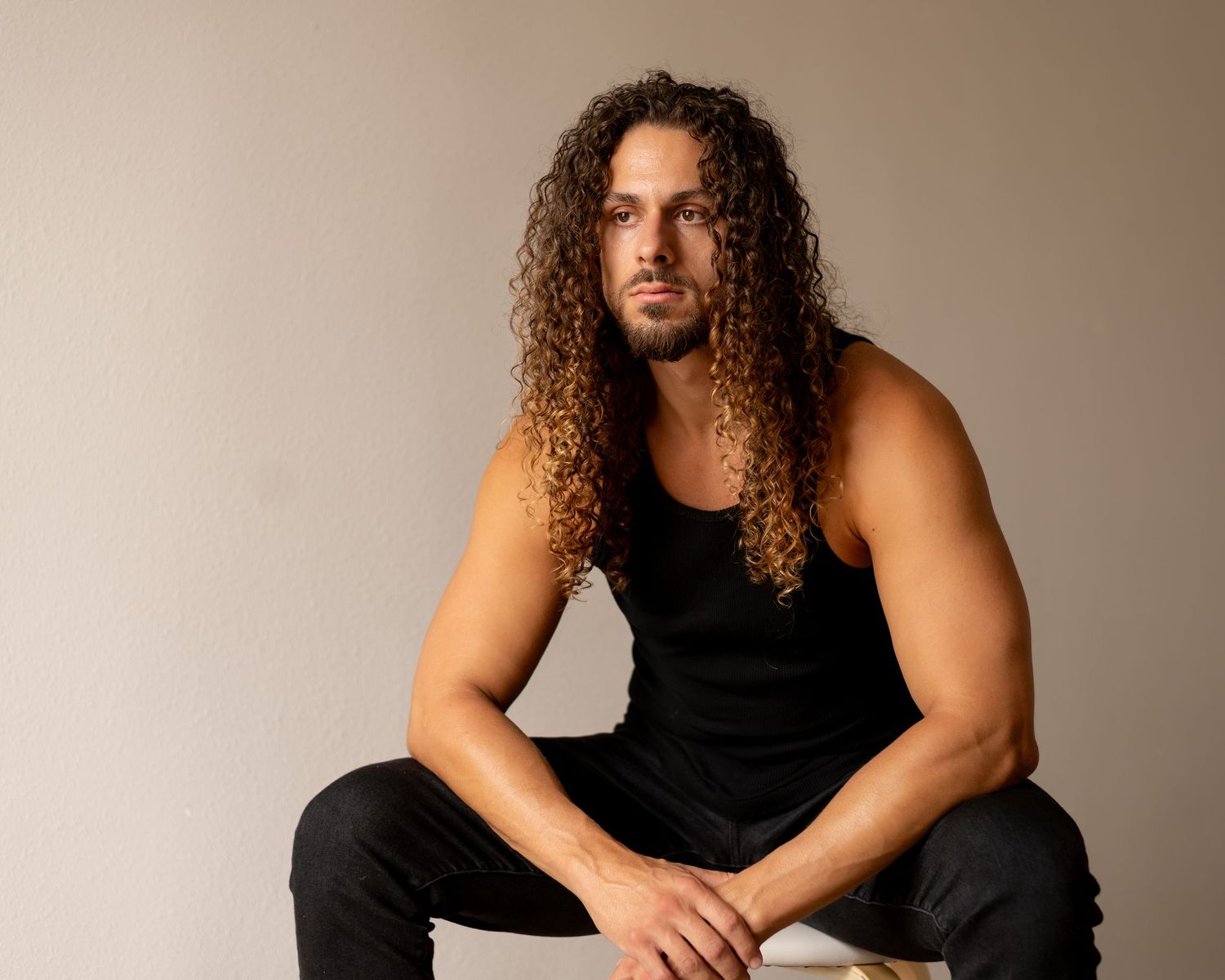 A person with long curly hair wearing a black tank top poses against a light colored backdrop in a photo series.