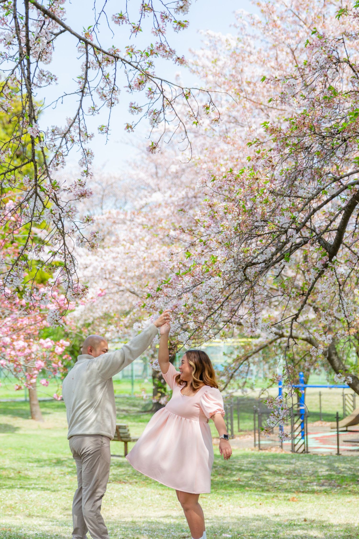 A couple dances playfully under blooming cherry blossom trees in a spring park setting.