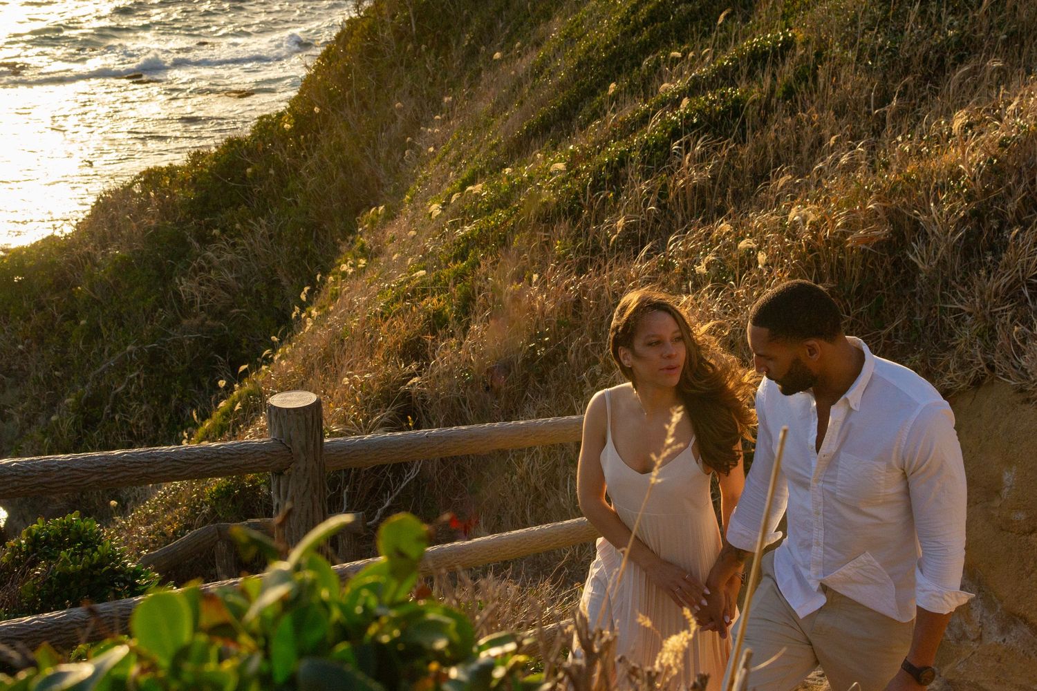 A couple walks hand in hand along a scenic coastal cliff path during golden hour.
