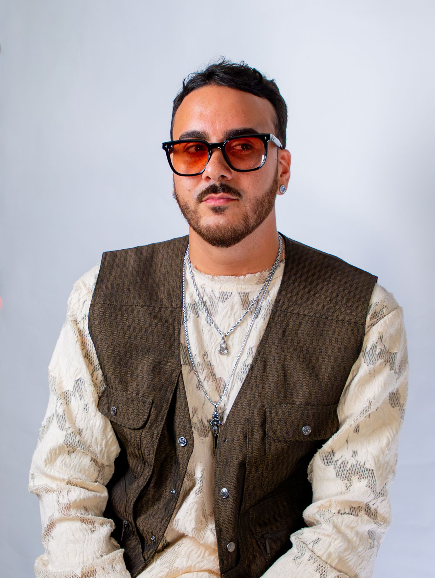 Person in brown vest and patterned shirt poses against white background wearing sunglasses.