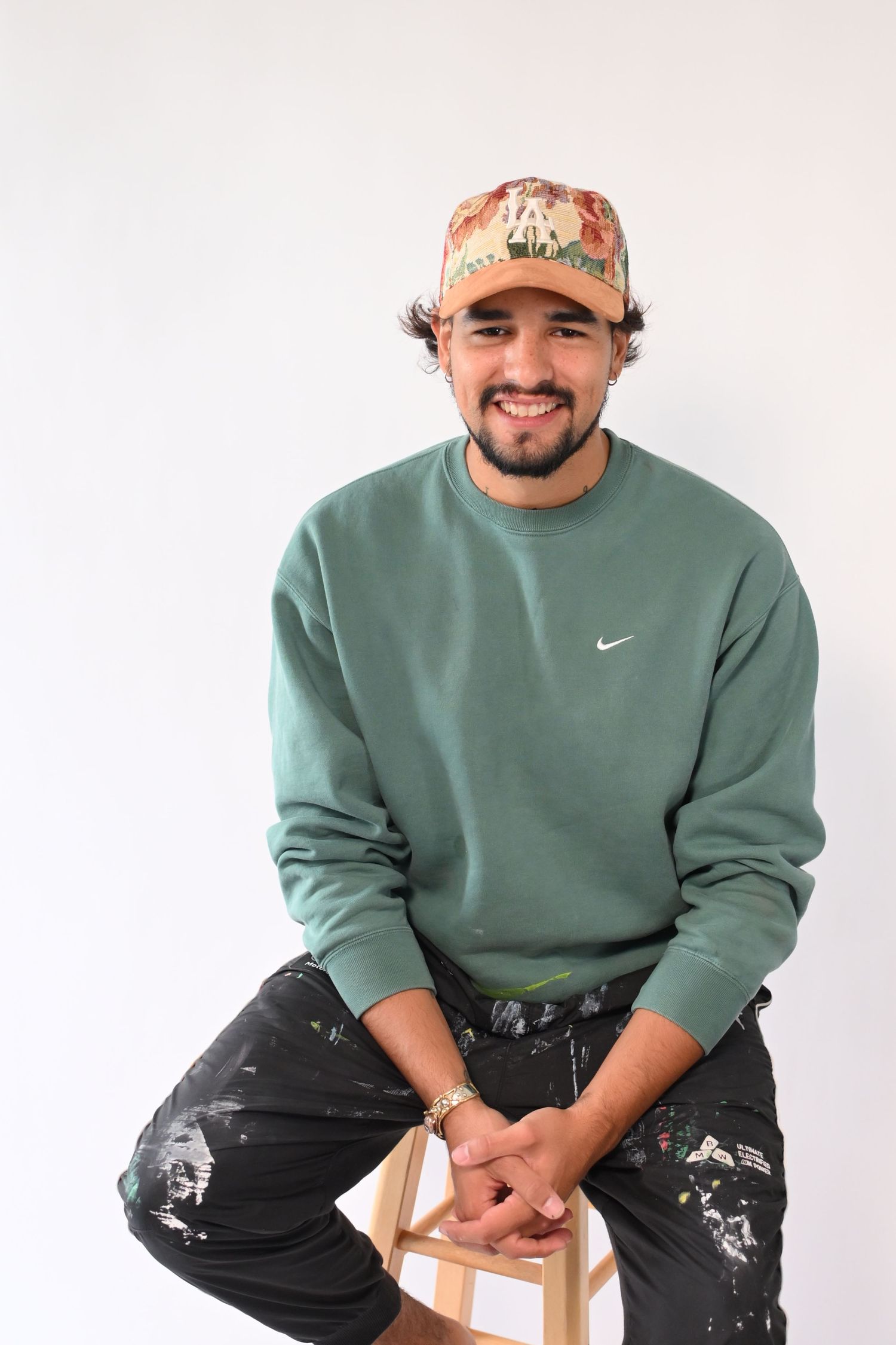 Person in sage green Nike sweatshirt and paint-splattered jeans sits casually on wooden stool against white background.