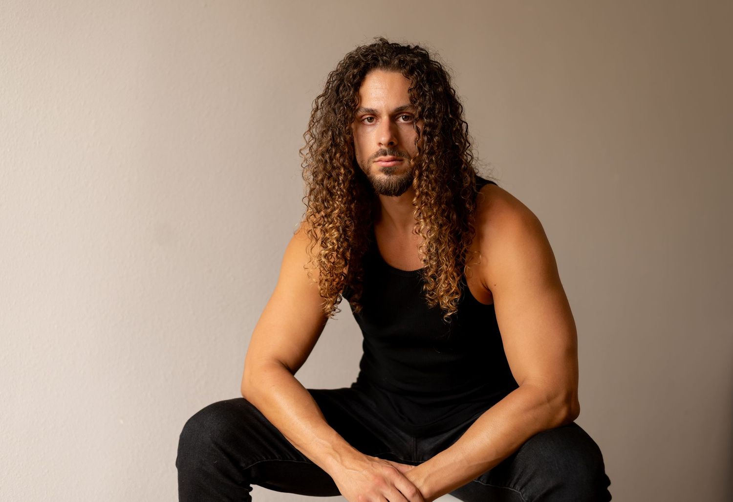 Studio portraits of a person with long curly hair wearing a black tank top against neutral background.