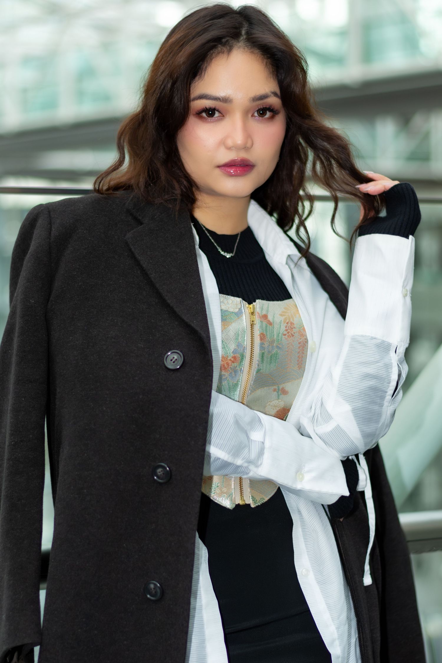 Professional wearing black coat and white blouse poses in modern office setting with glass windows.