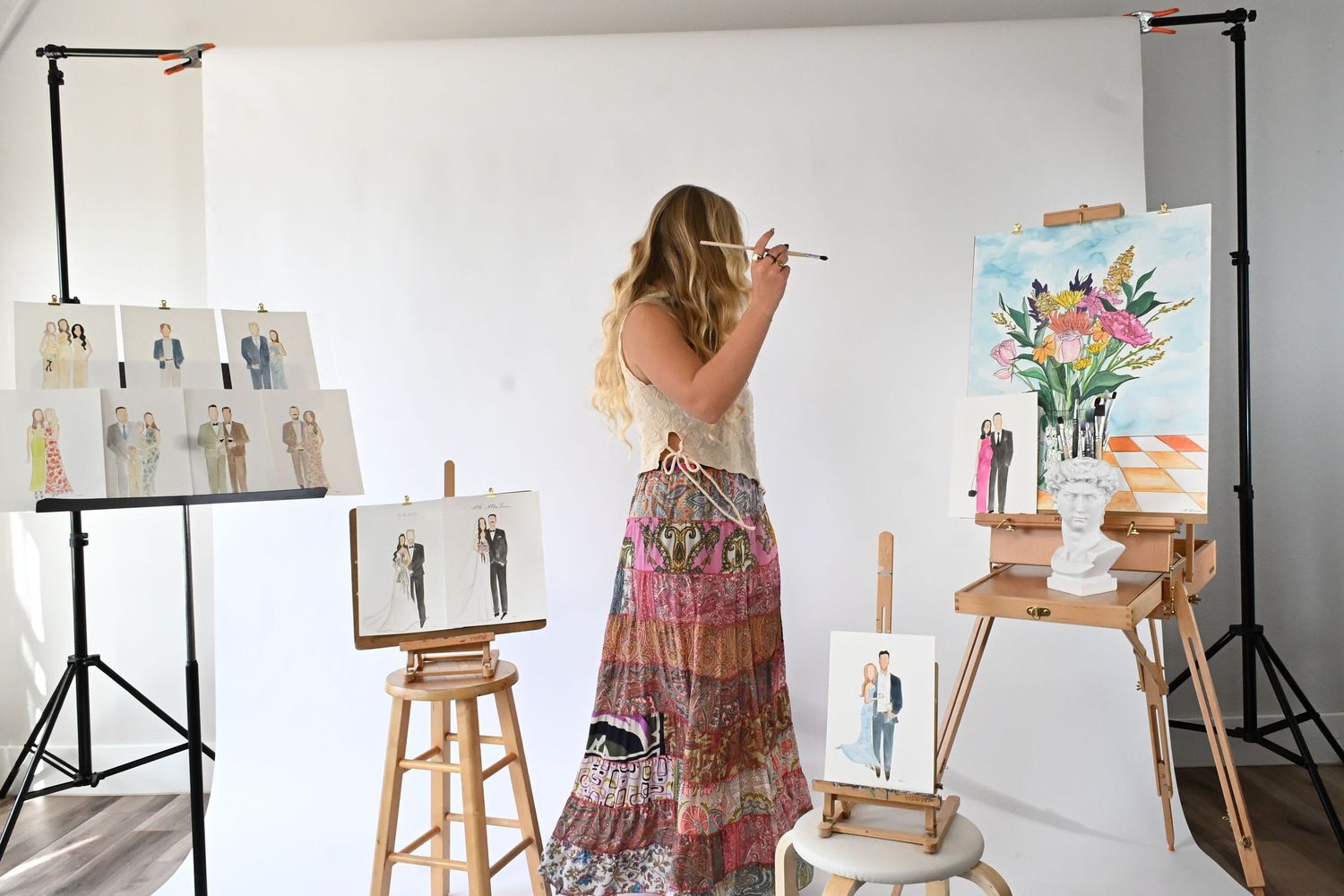 Artist working at easel in bright studio space with multiple paintings and sketches displayed on white walls.
