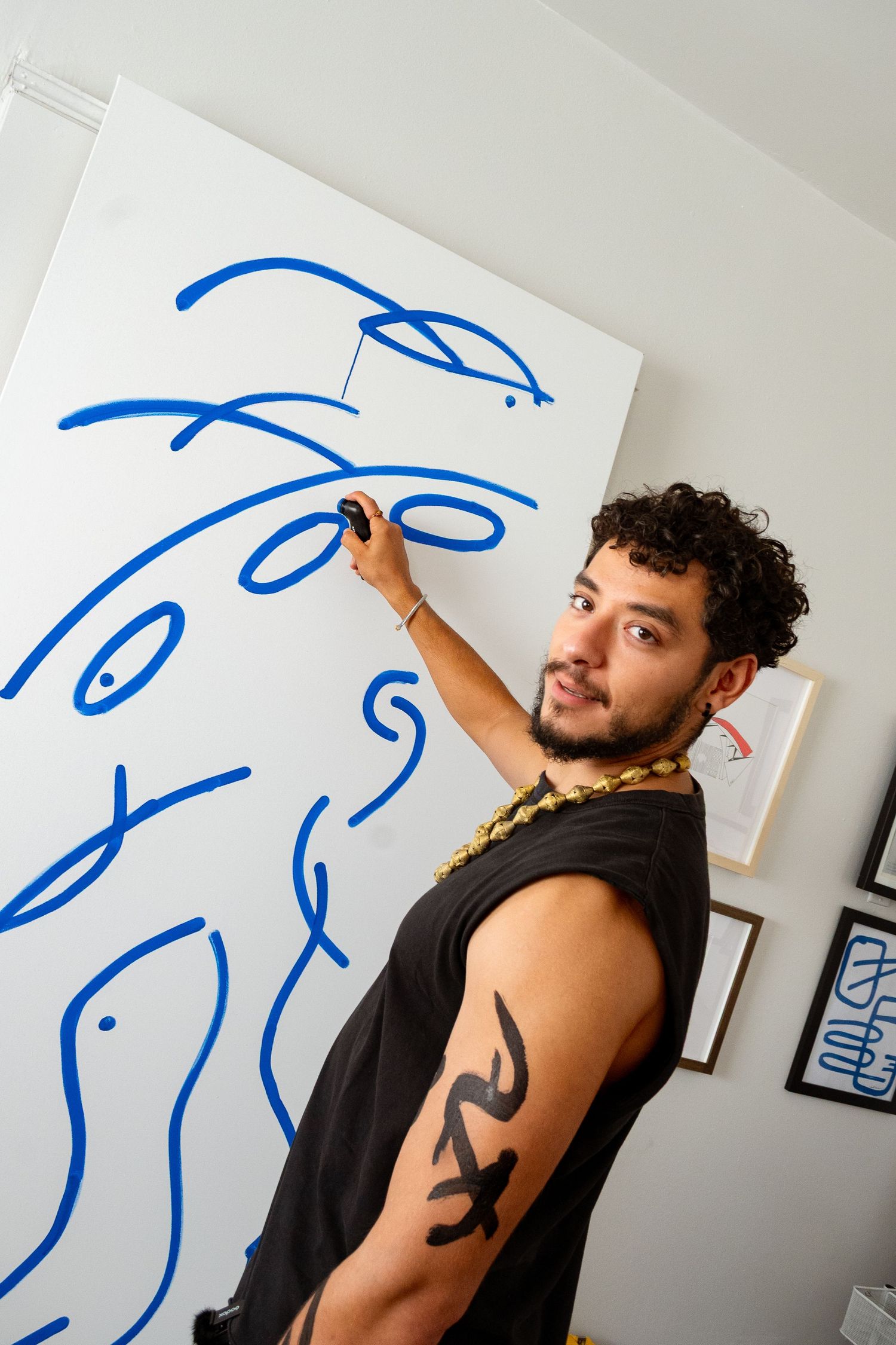 Artist draws flowing blue line art on white paper mounted to wall in progressive sequence.