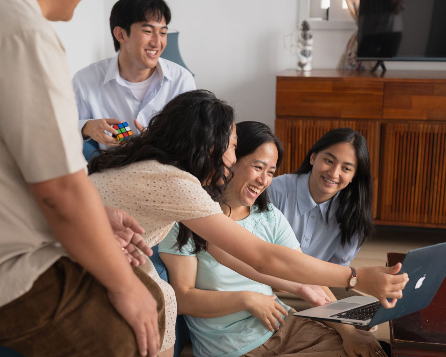 A group of smiling Asian friends gather around a laptop together in a cozy living room setting.