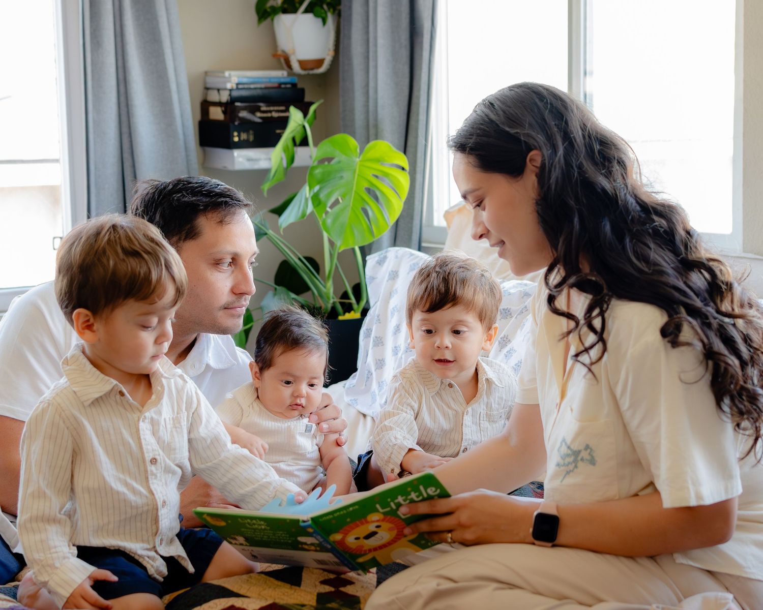 Family sitting together on couch reading a children's book with green houseplant in background.