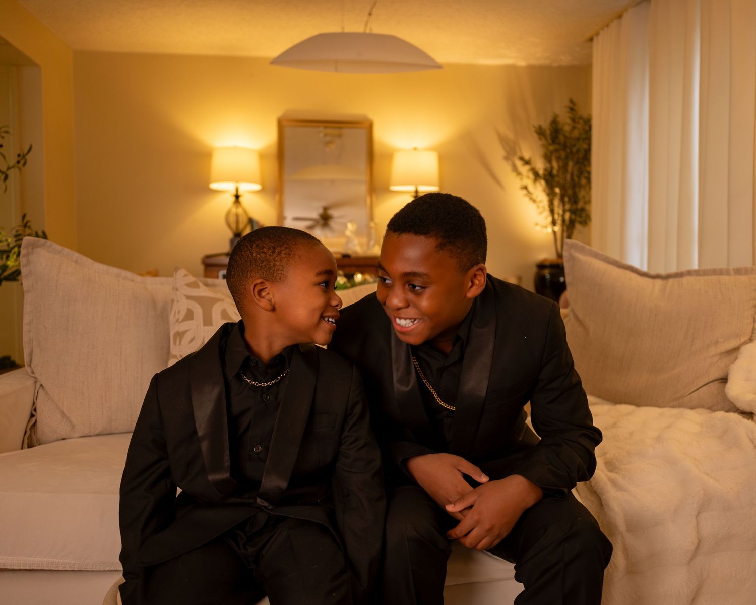 Two young boys in matching black suits sit together on a white couch, smiling at each other in a warmly lit room.