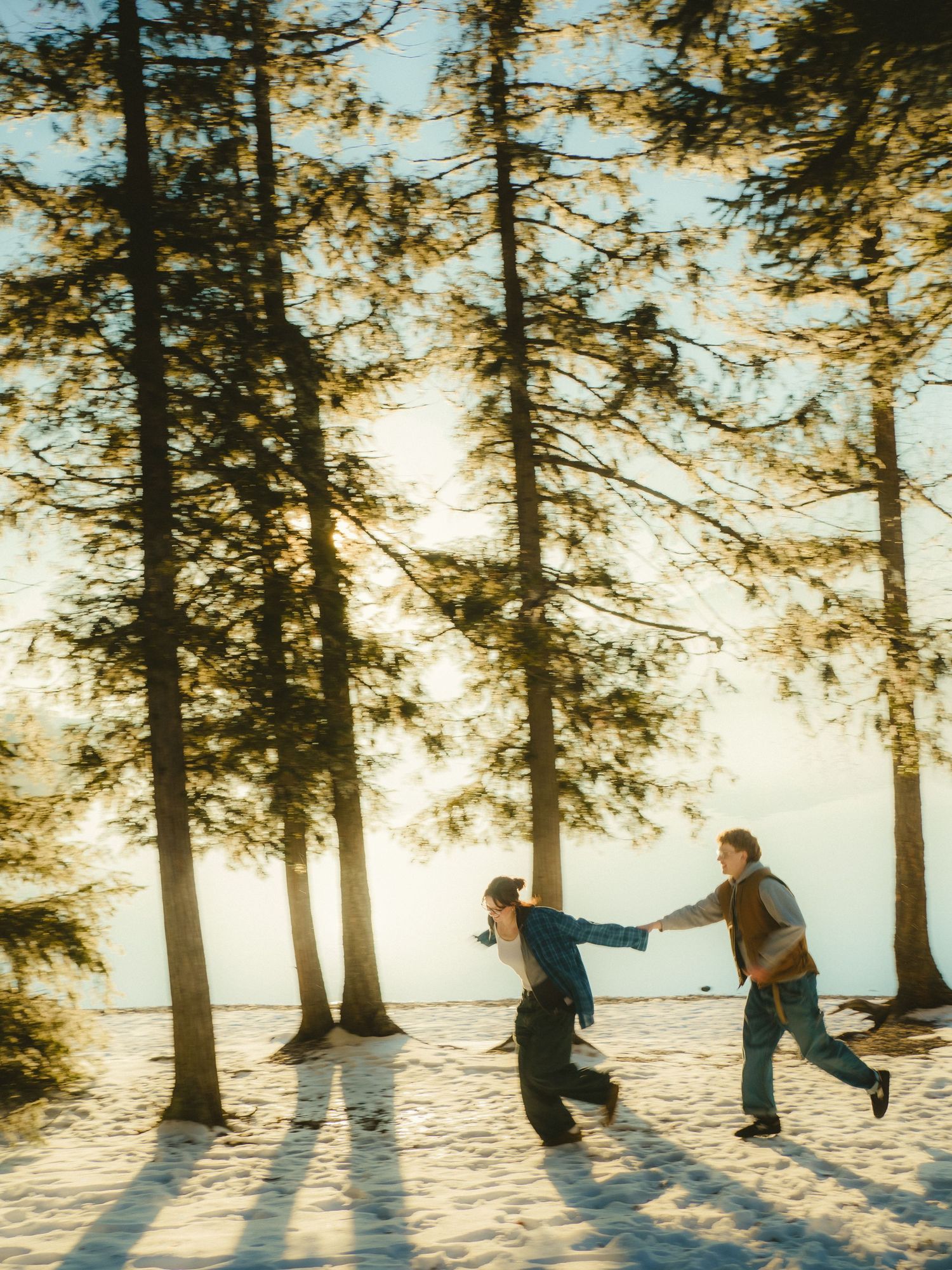 Couple hold hands and run through the snow and pine trees with Lake McDonald behind them in Glacier National Park, Montana.