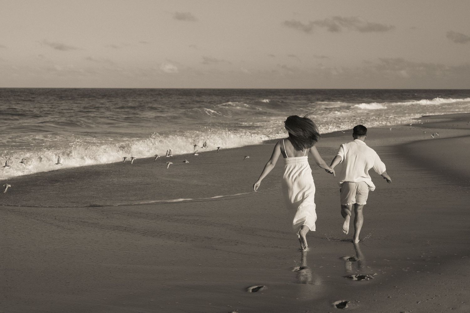 Couple holding hands while running down the beach as seagulls fly past in Dewey Beach, Delaware.