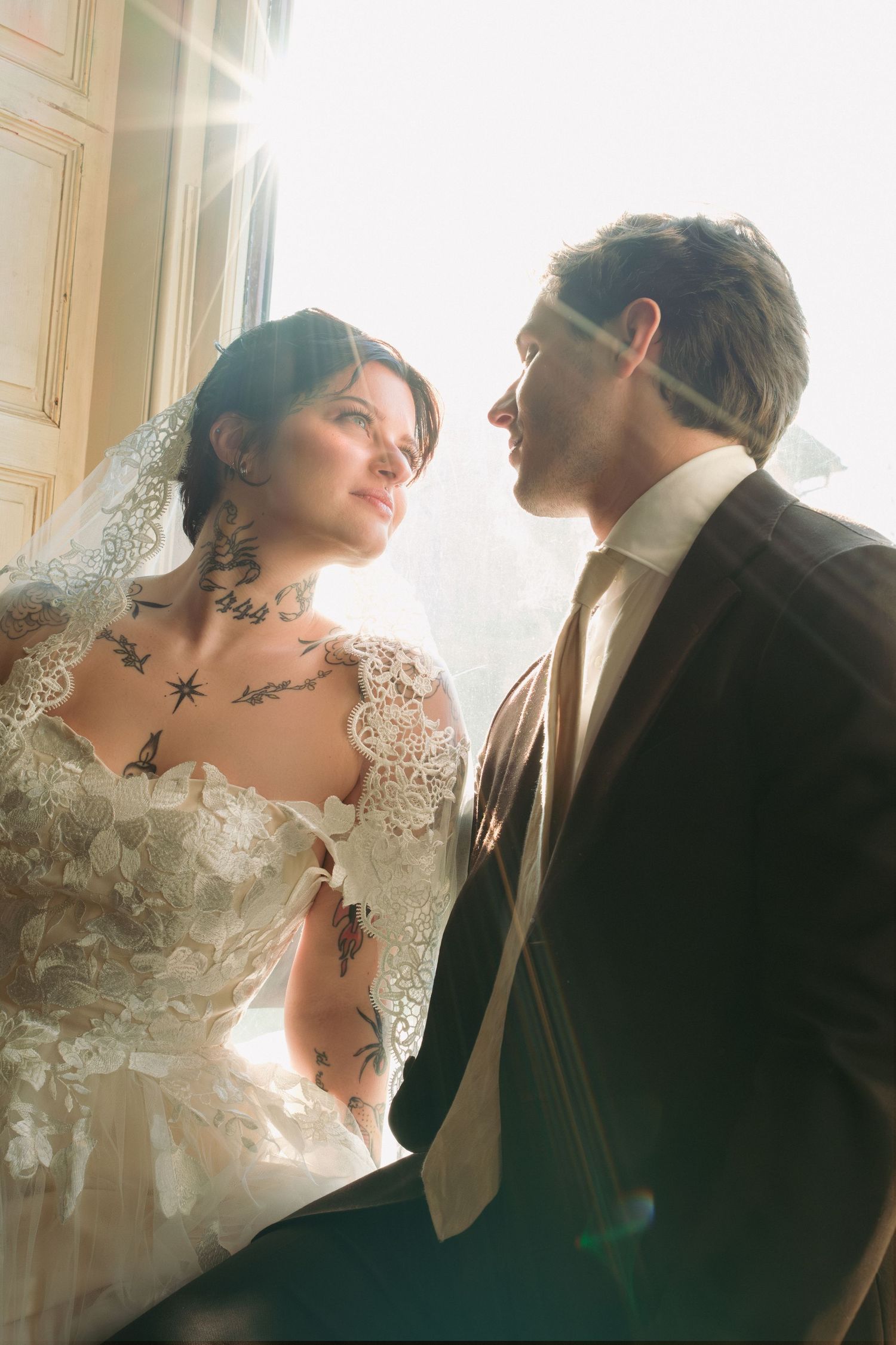 Bride with tattoos in lace wedding dress and veil gazes at groom in suit, bathed in sunlight by a window.