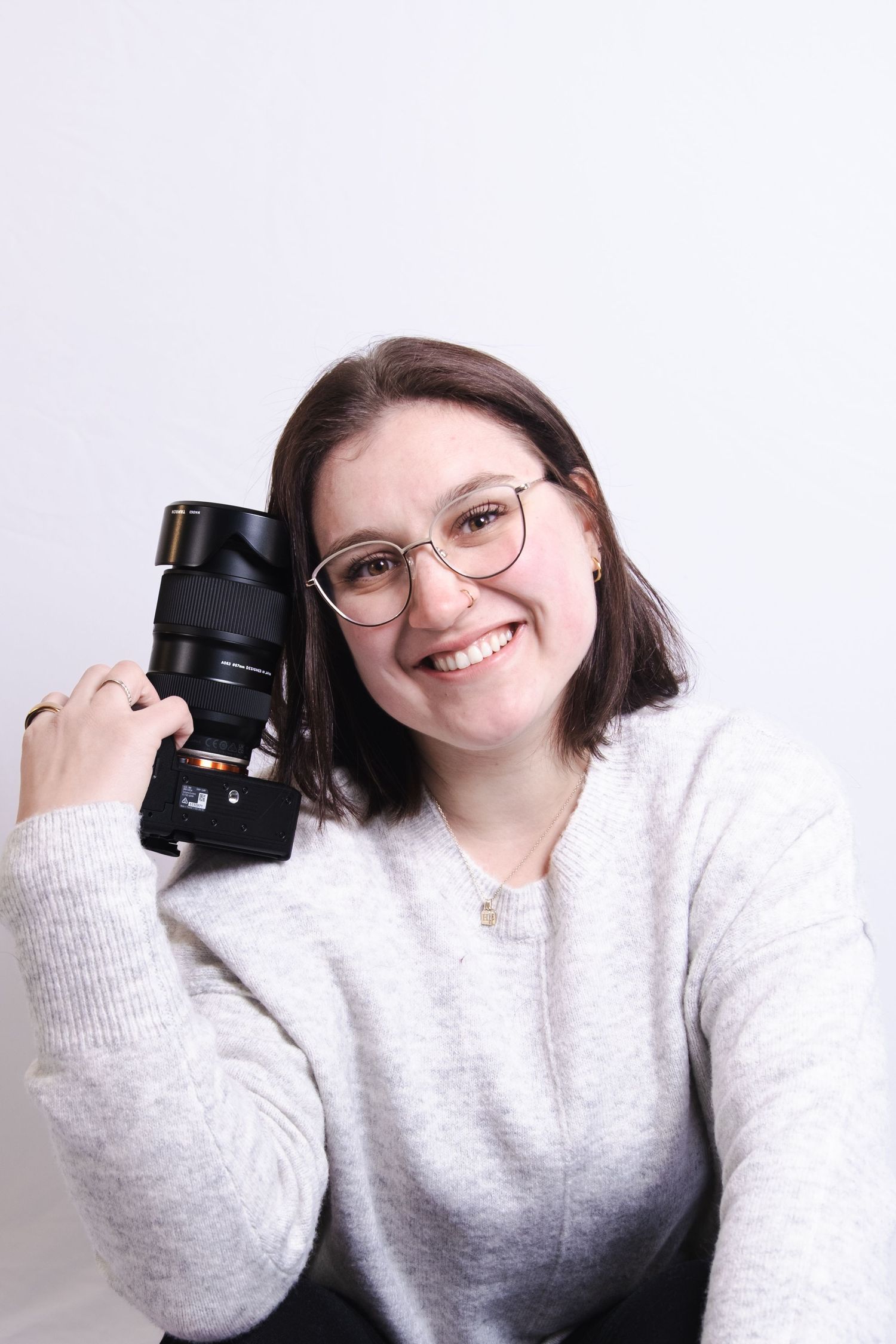 A photographer in a light gray sweater holds up a camera lens while posing for multiple playful shots.