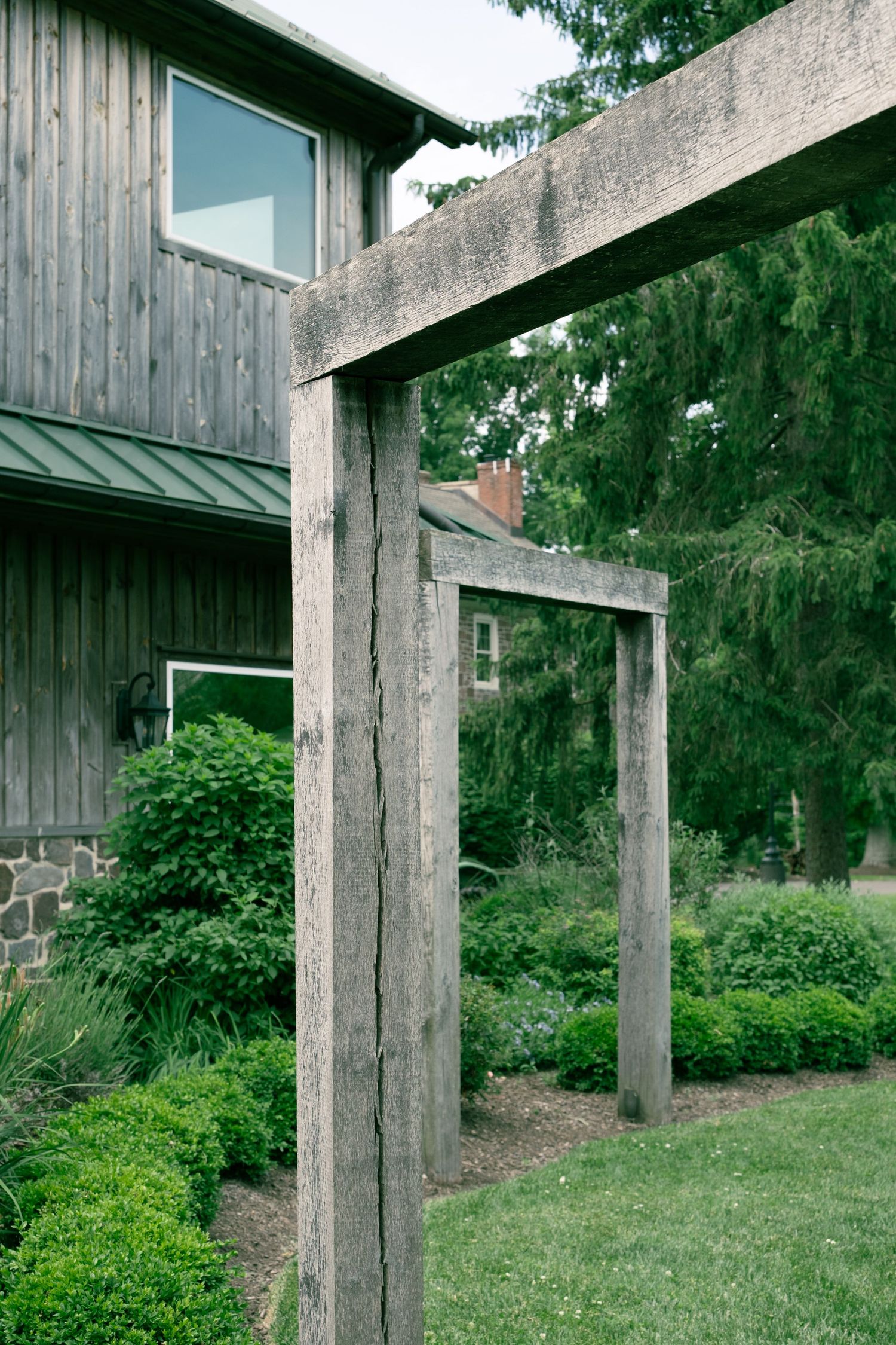 Rustic wooden pergola beside a weathered barn with stone foundation, surrounded by lush green gardens and manicured shrubs.