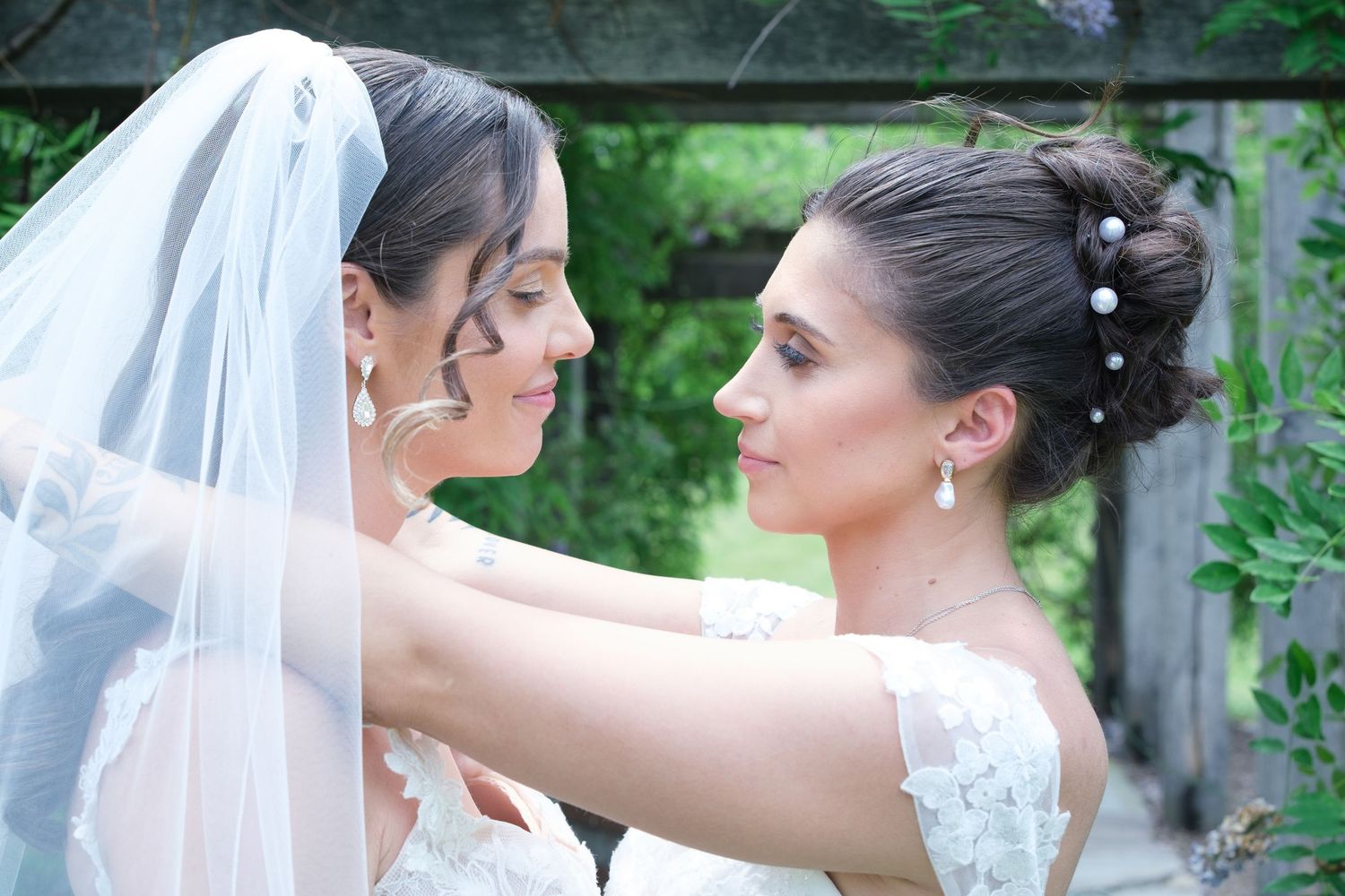 Two brides in white lace wedding gowns face each other tenderly outdoors, one wearing a veil.
