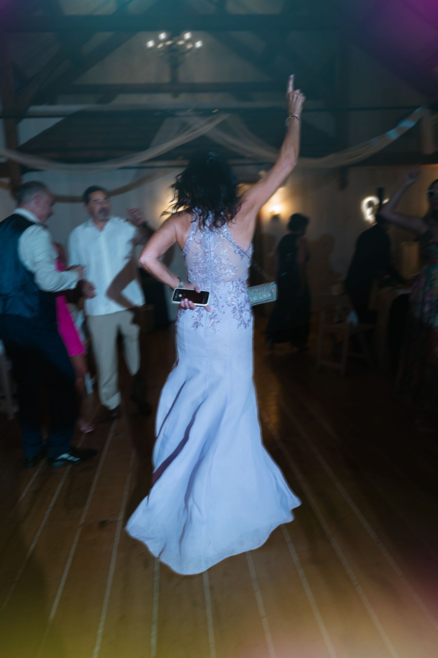 Bride in white mermaid gown dancing with arm raised at lively wedding reception on wooden dance floor.