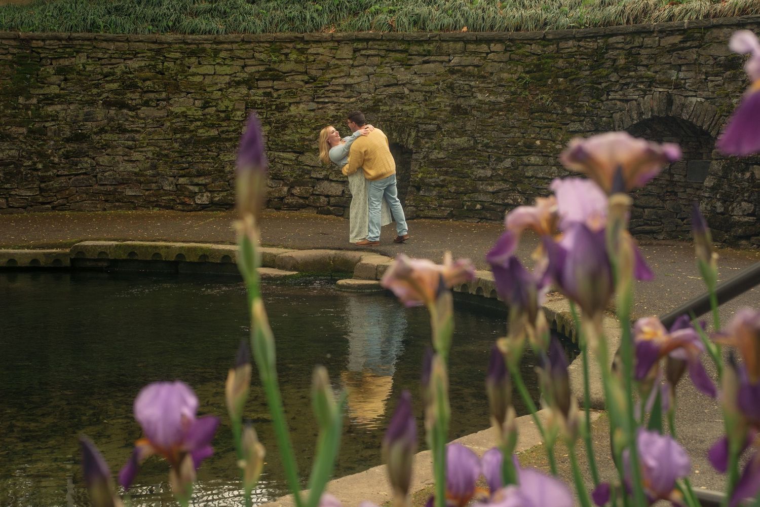 Couple embracing near a tranquil garden pond surrounded by blooming purple iris flowers.