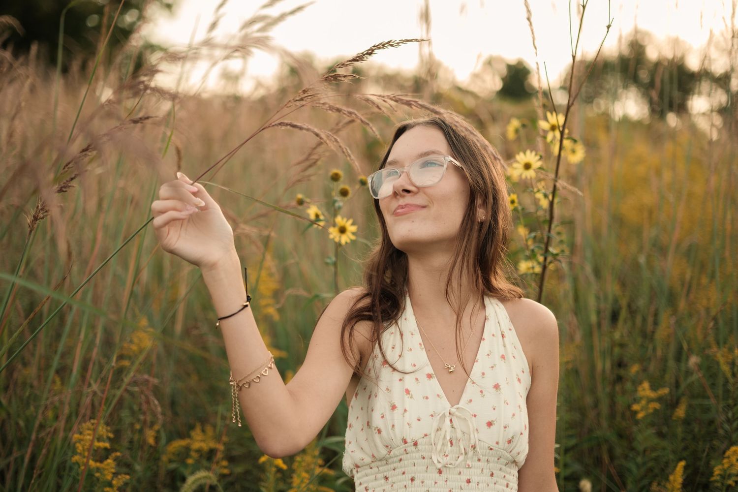 Young woman in floral dress and glasses standing in a wildflower field at golden hour, reaching up to touch tall grass.