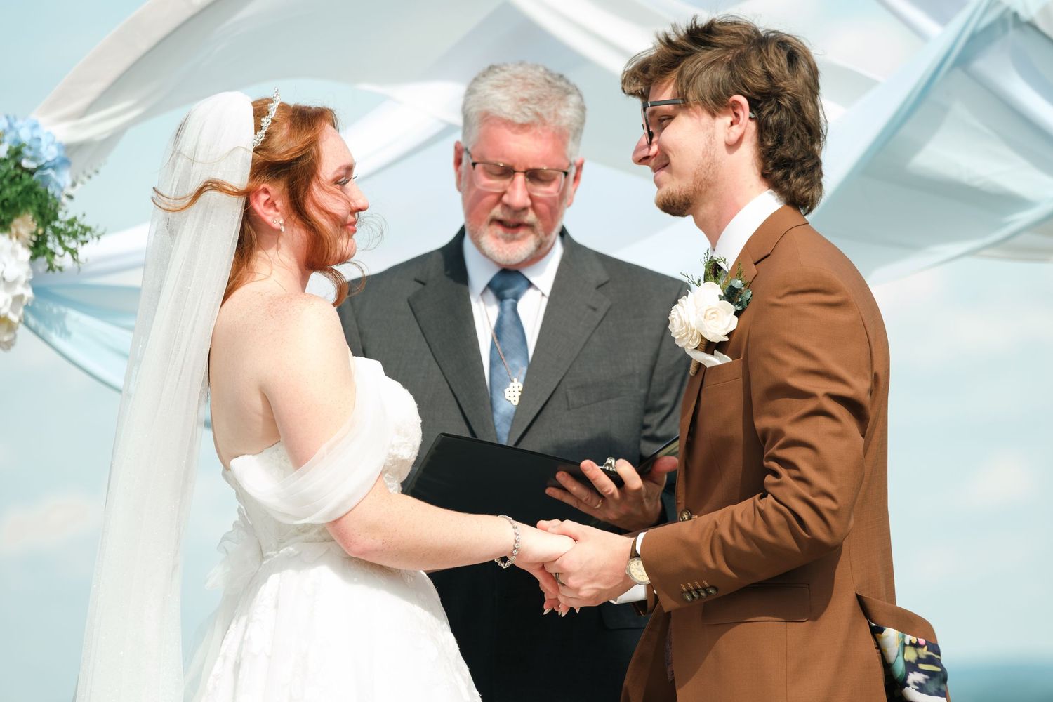 Bride and groom exchange vows at outdoor wedding ceremony with officiant holding black book.