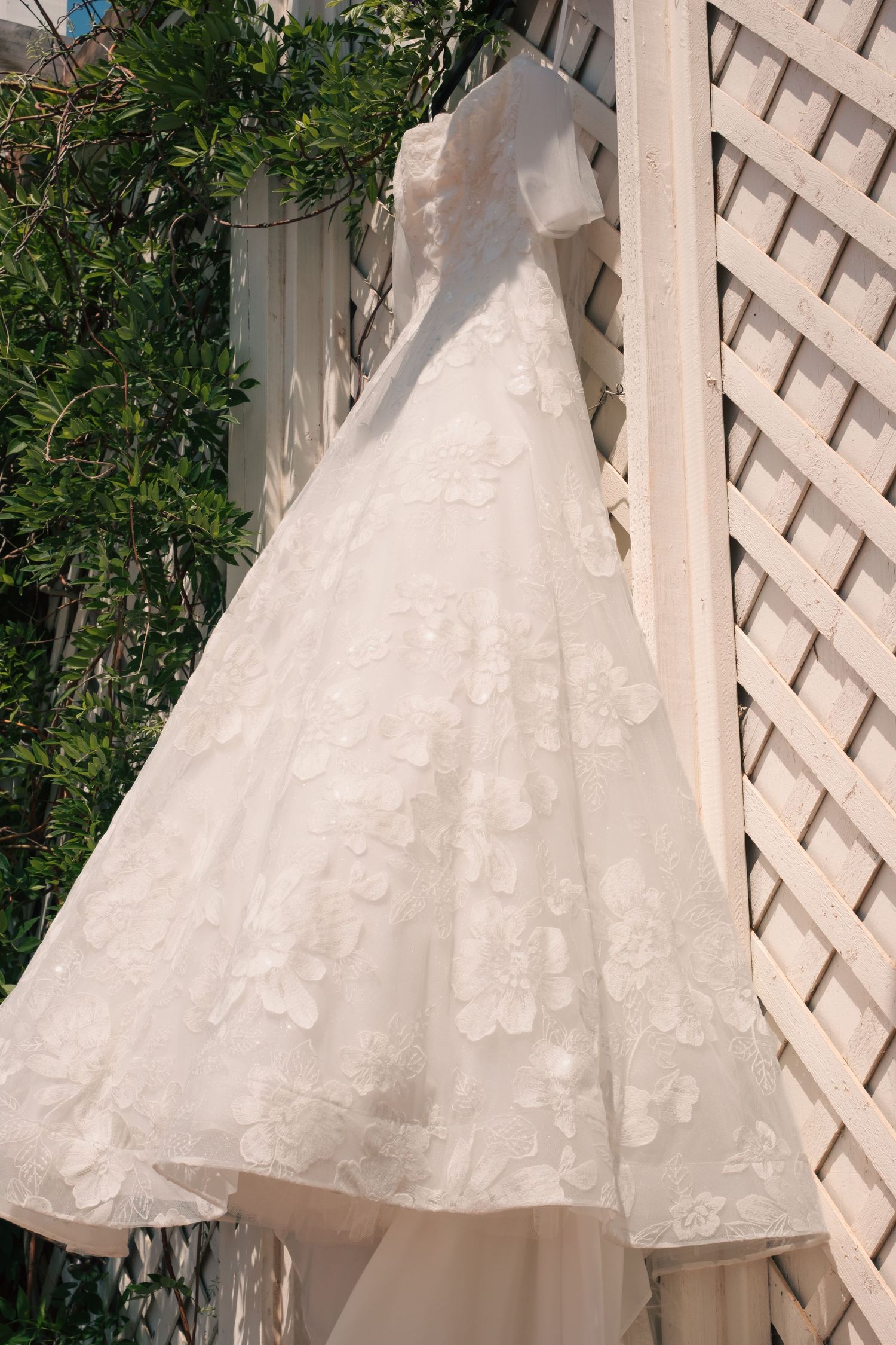 Elegant white lace wedding dress with floral appliques hanging on a wooden lattice outdoors.