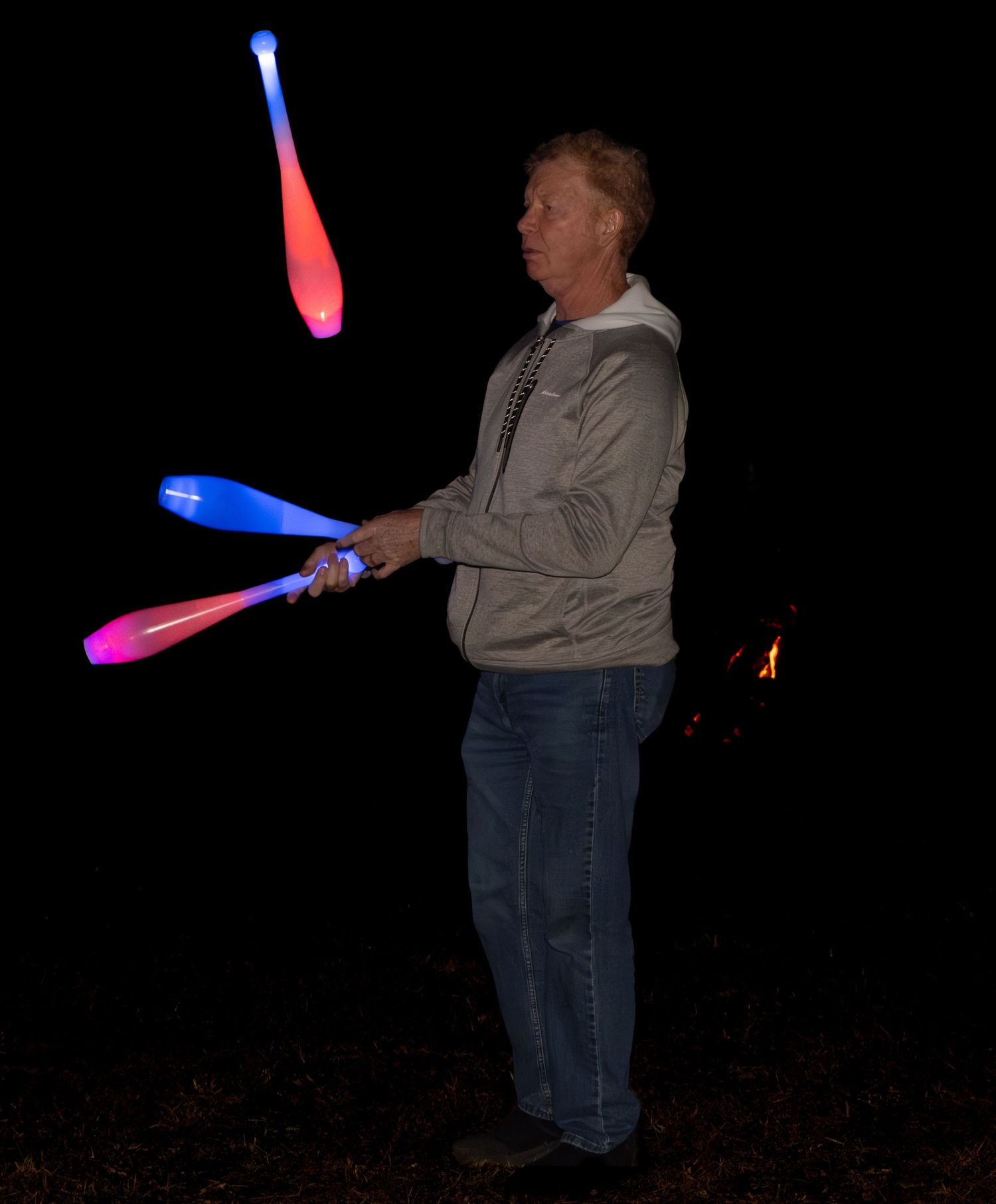 Person juggling illuminated LED clubs in a dark outdoor setting creating colorful light trails in the night.