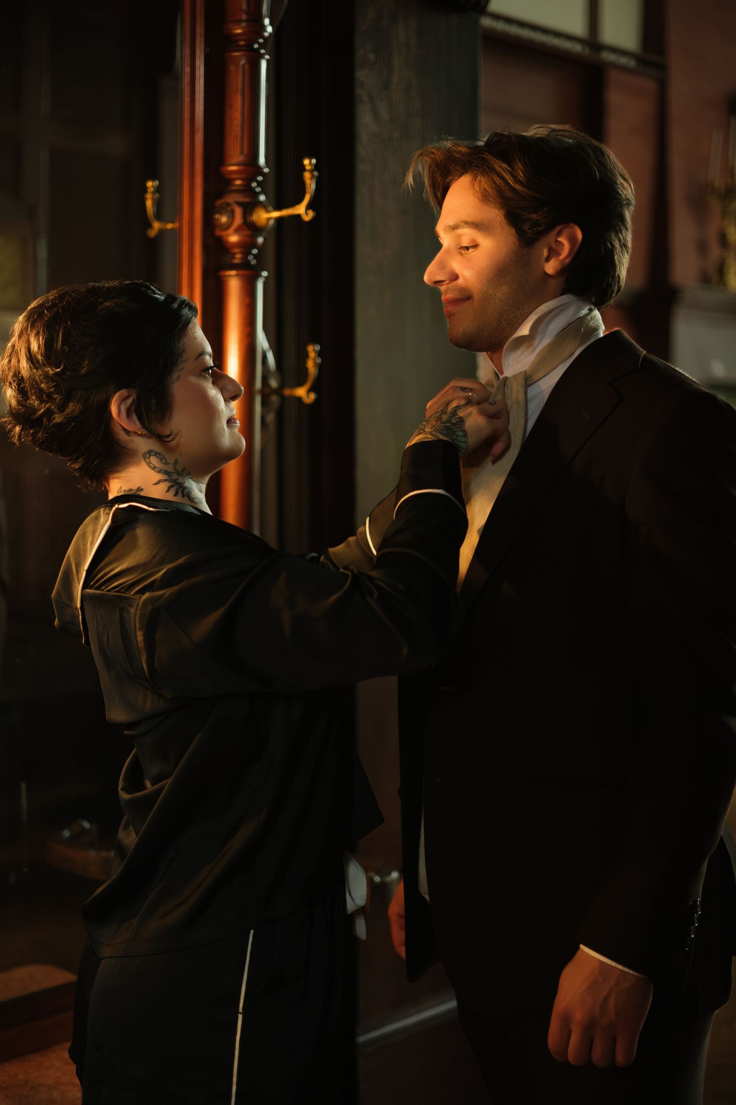 A woman adjusts a man's collar in a dimly lit, vintage-styled room, both dressed in elegant attire.
