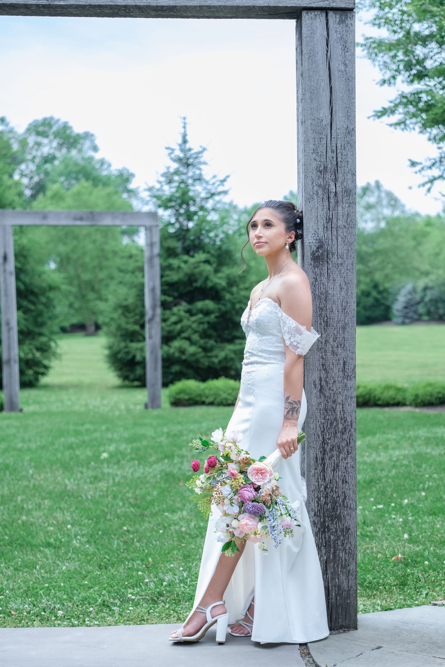 Bride in white off-shoulder gown holding colorful bouquet, leaning against wooden post in lush green garden setting.