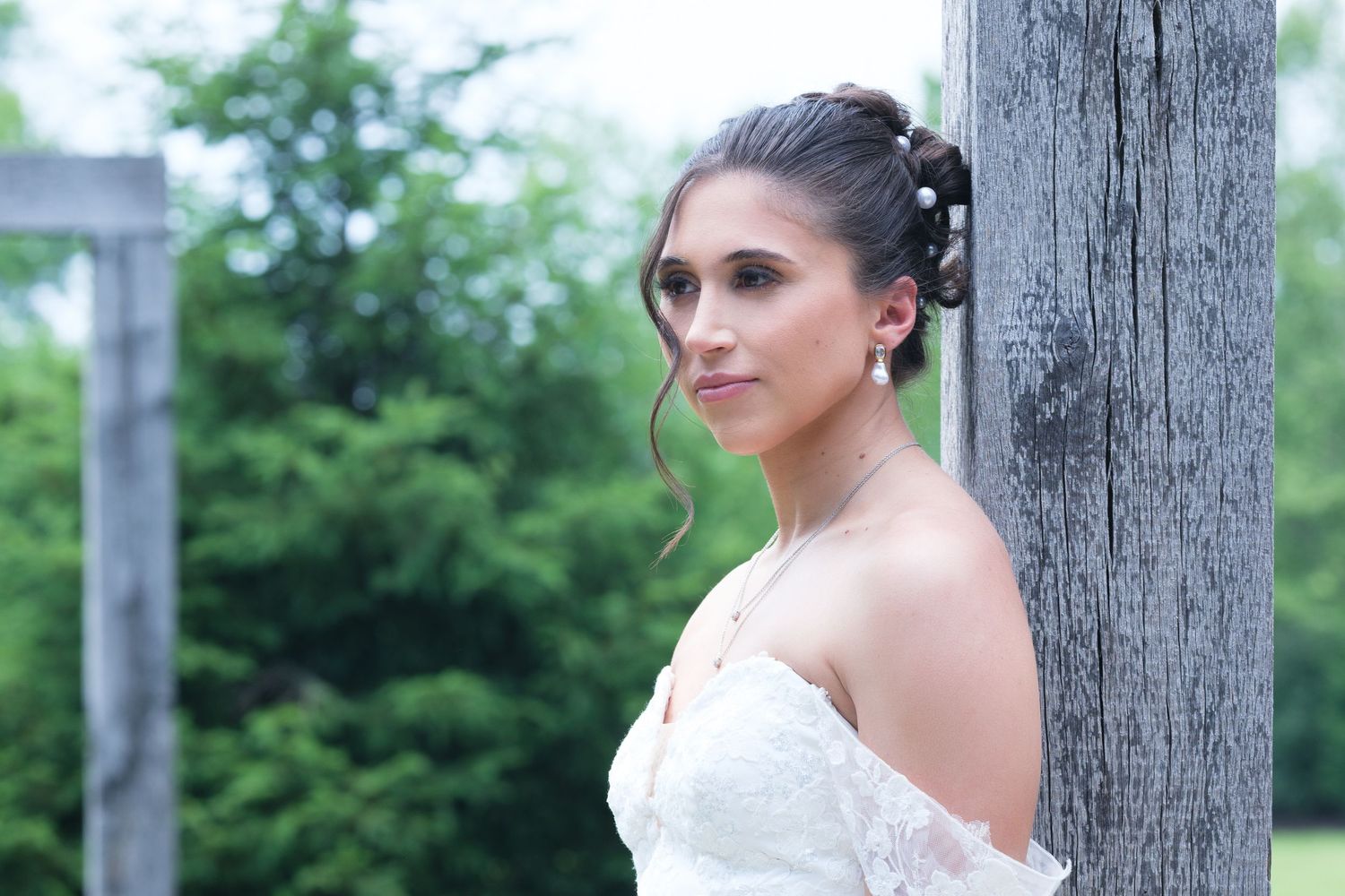 Bride in white strapless gown poses gracefully beside rustic wooden post with lush green trees in background.