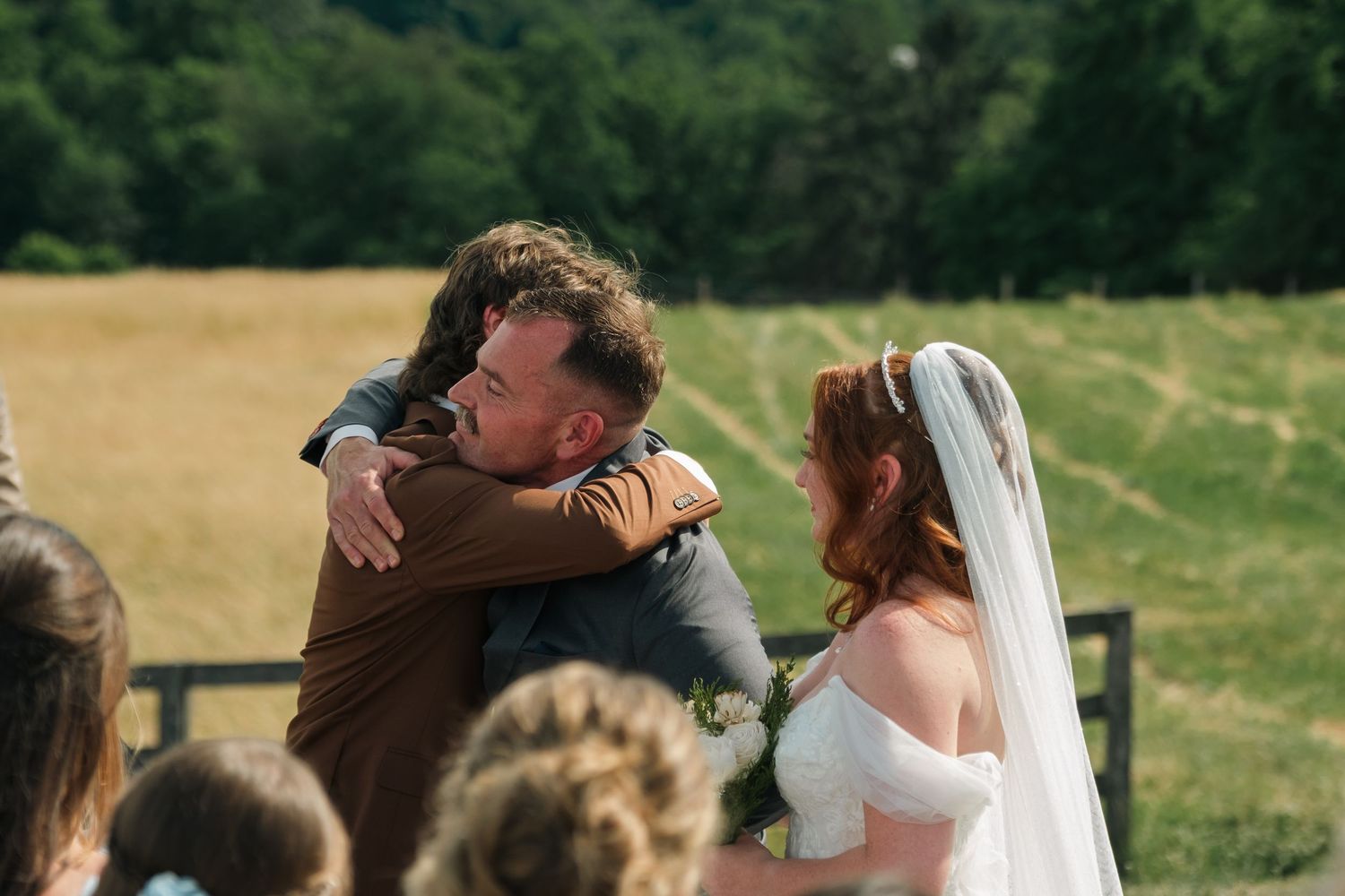 Groom embracing a guest while bride in white dress and veil smiles during outdoor wedding ceremony.