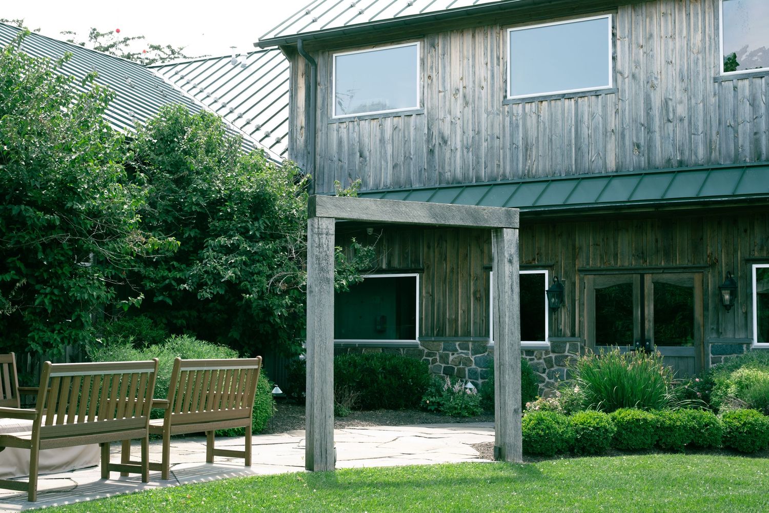 Rustic wooden barn venue with green metal roof, stone patio, and garden benches surrounded by lush landscaping.