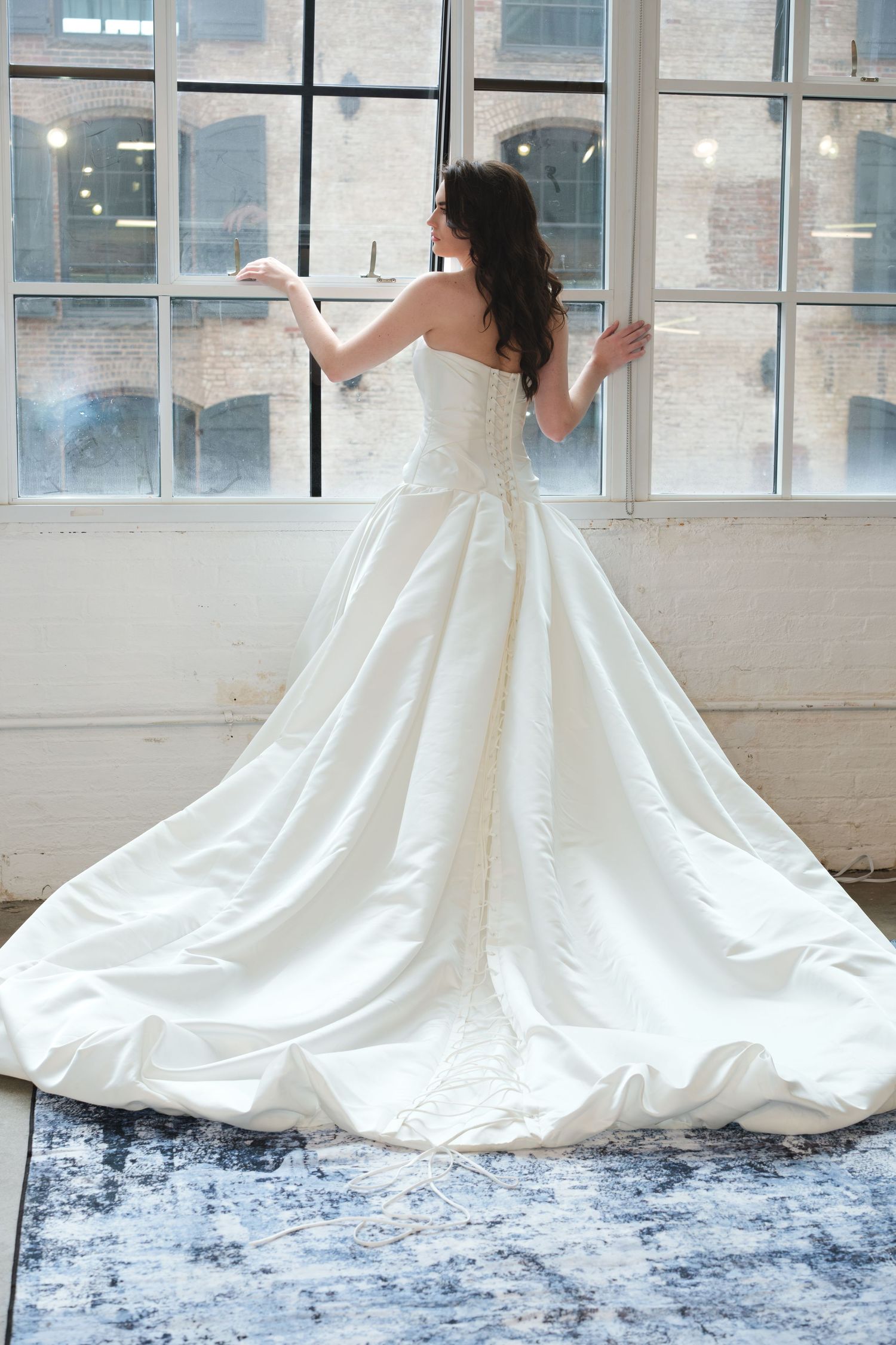 Bride in strapless white ballgown with long train stands before large windows in bright studio.