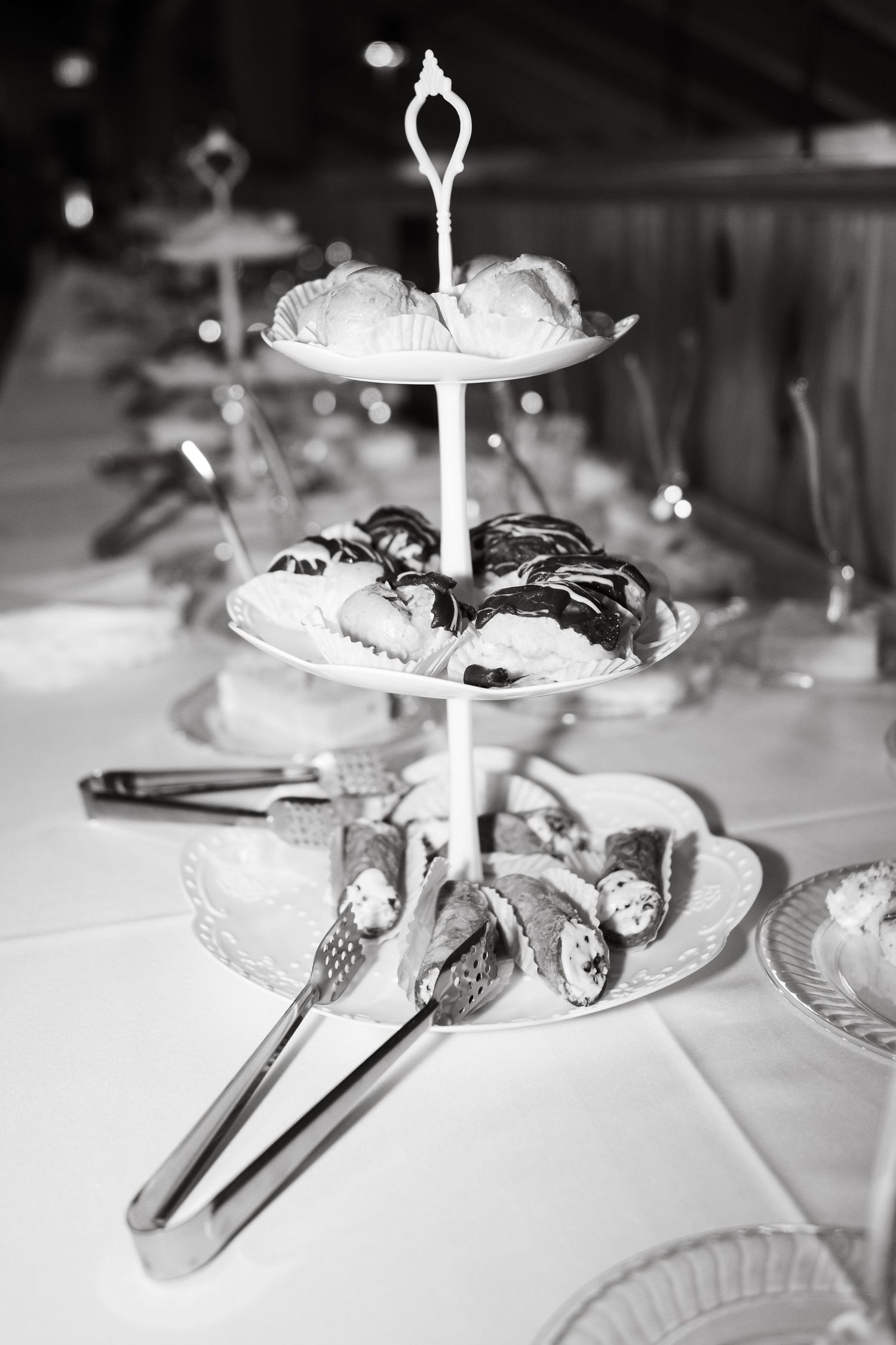 Black and white photo of a tiered dessert stand with pastries and tongs on a white tablecloth at an elegant event.