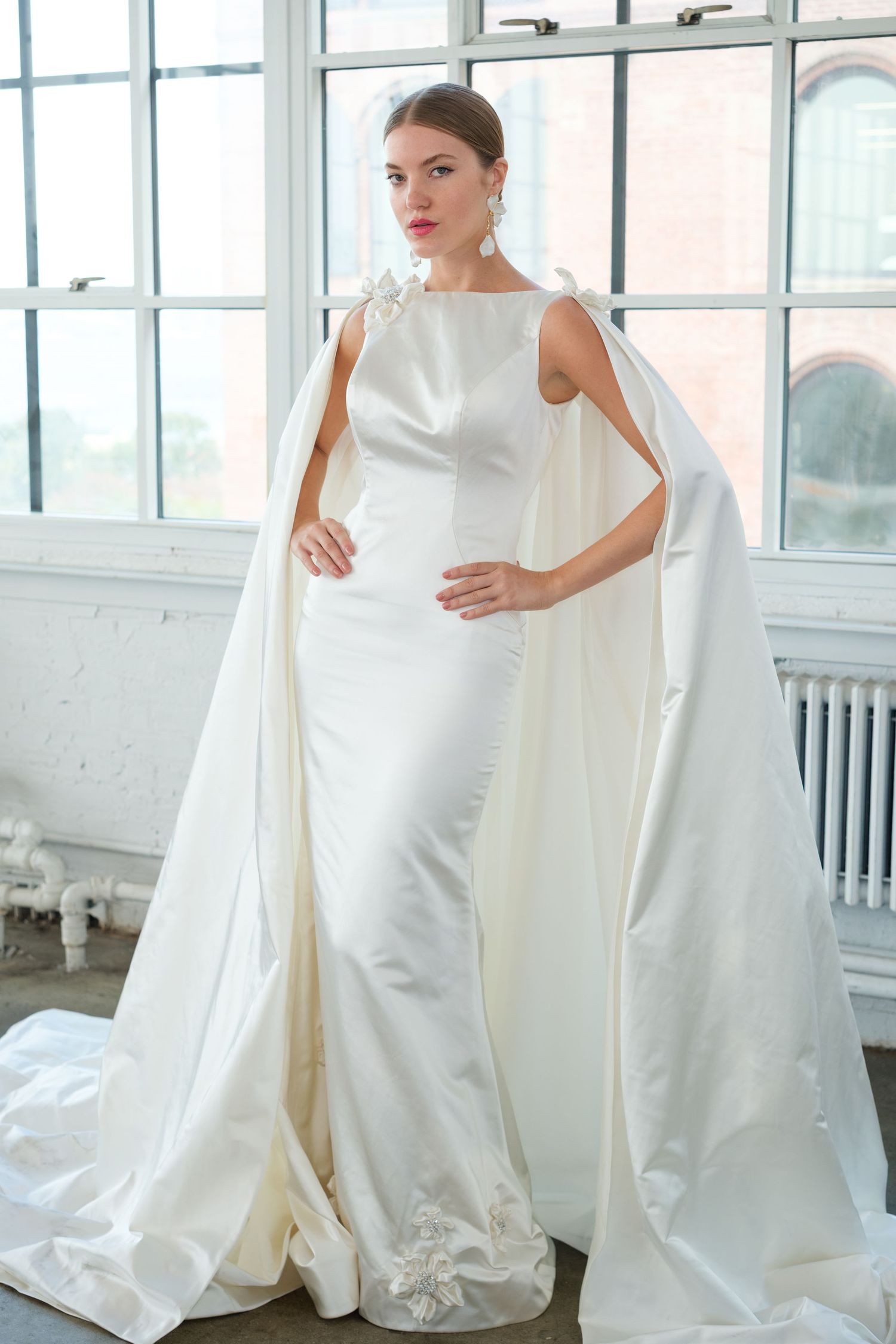 Elegant bride in a sleeveless white satin wedding gown with a dramatic floor-length cape and floral details.