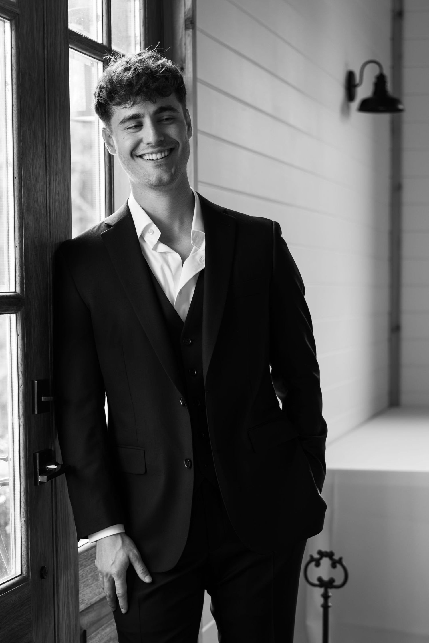 Smiling young man in black suit and open-collar shirt leaning against wall near window in black and white photo.