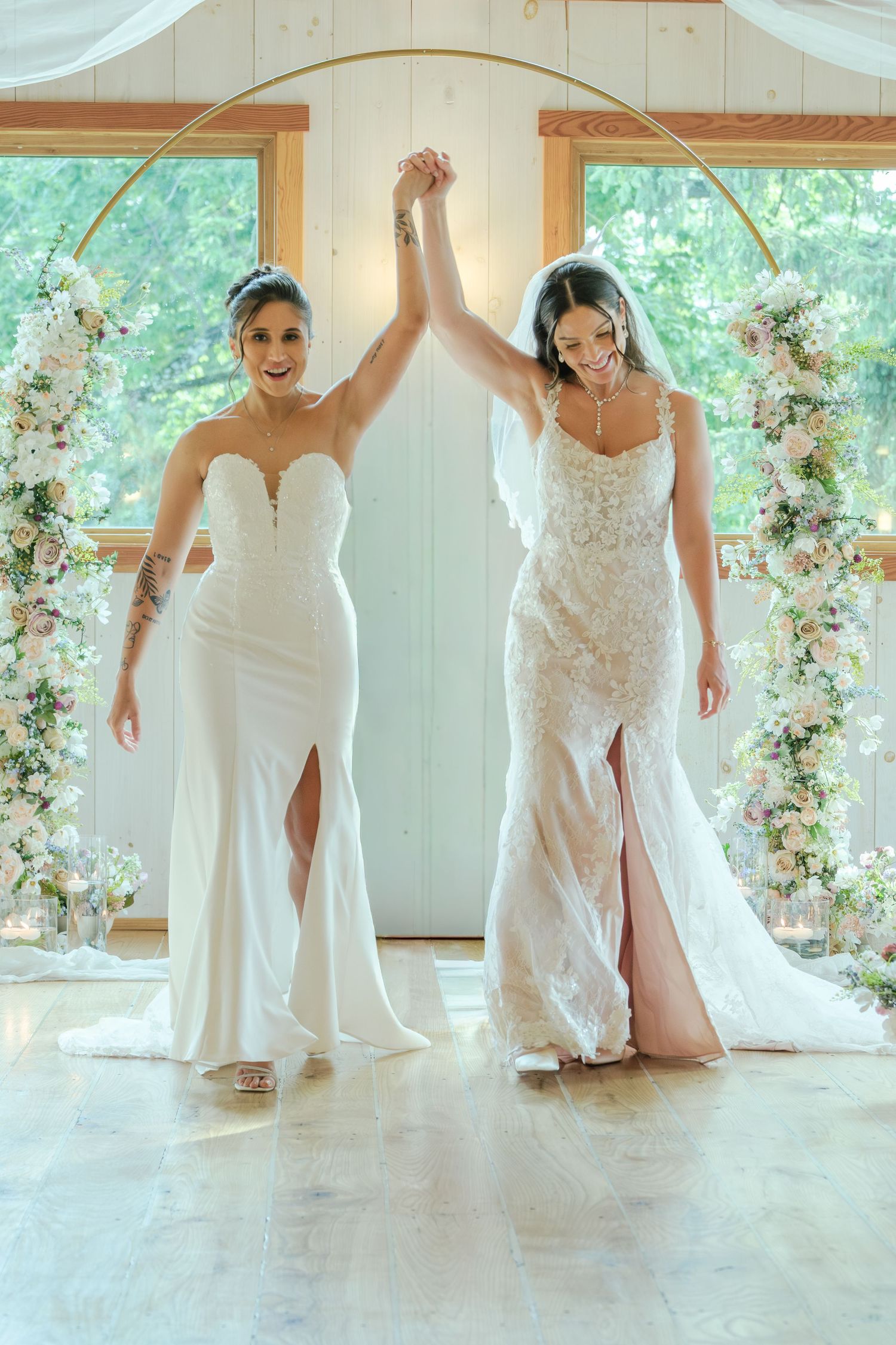 Two brides in white wedding gowns raise hands together under a floral arch at their wedding ceremony.