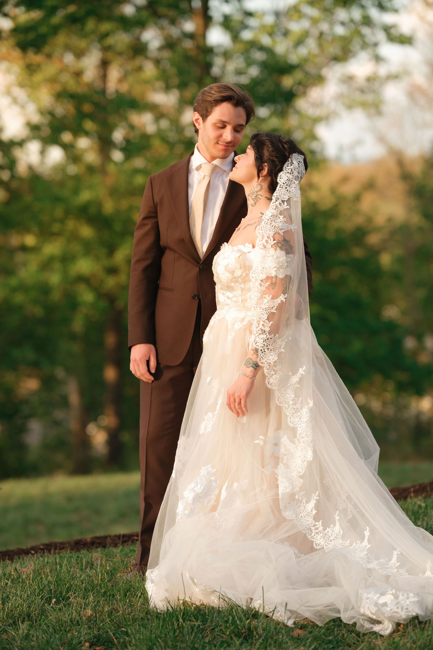 Bride in lace wedding dress and veil stands with groom in brown suit outdoors in a sunlit green park.