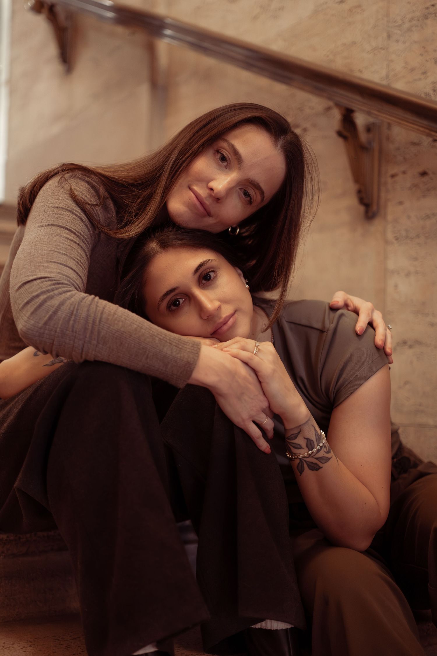 Two people in gray and brown clothing sit close together in a dimly lit room with wooden details.