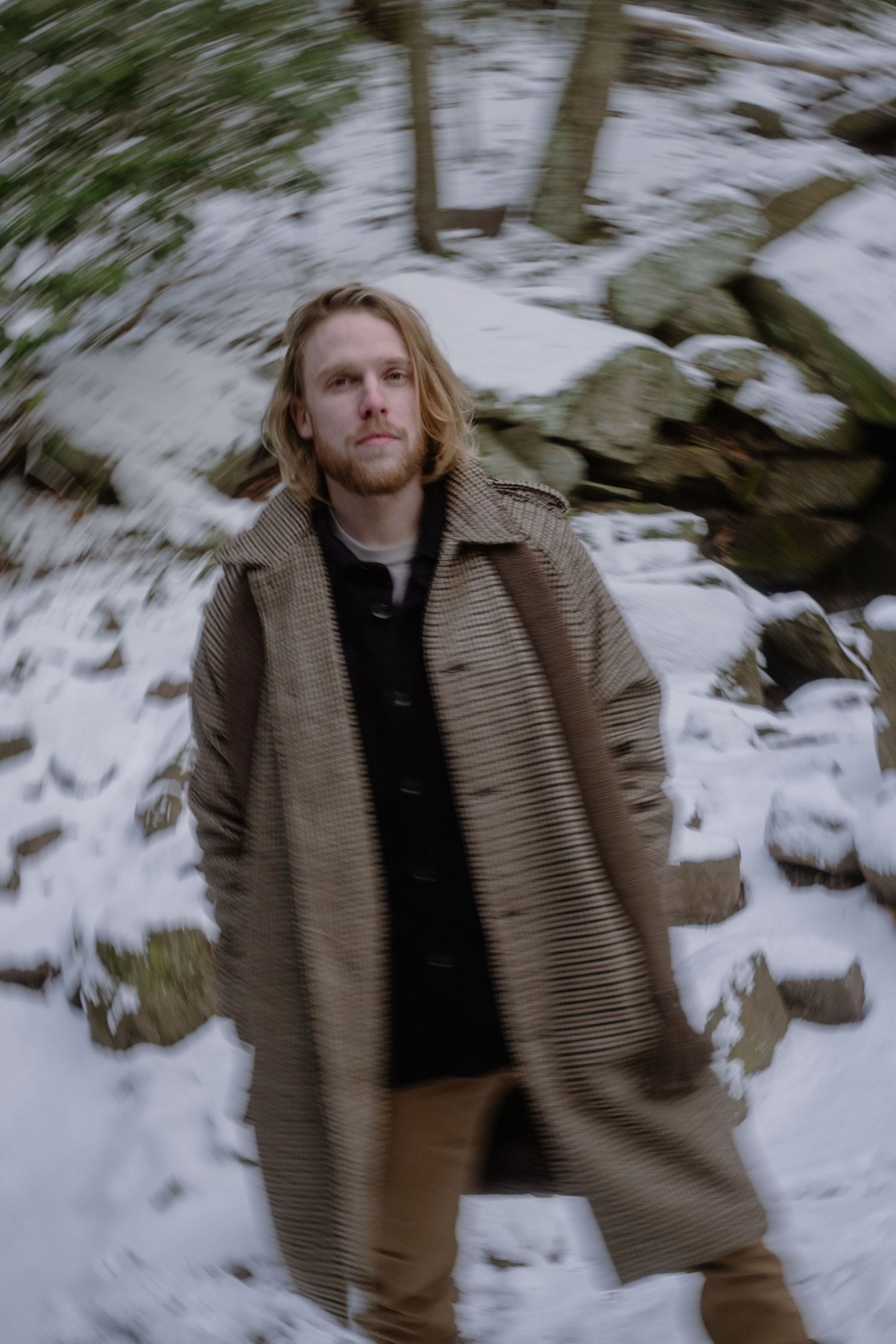 A person in a brown coat walks through a snowy winter landscape with blurred motion effects.