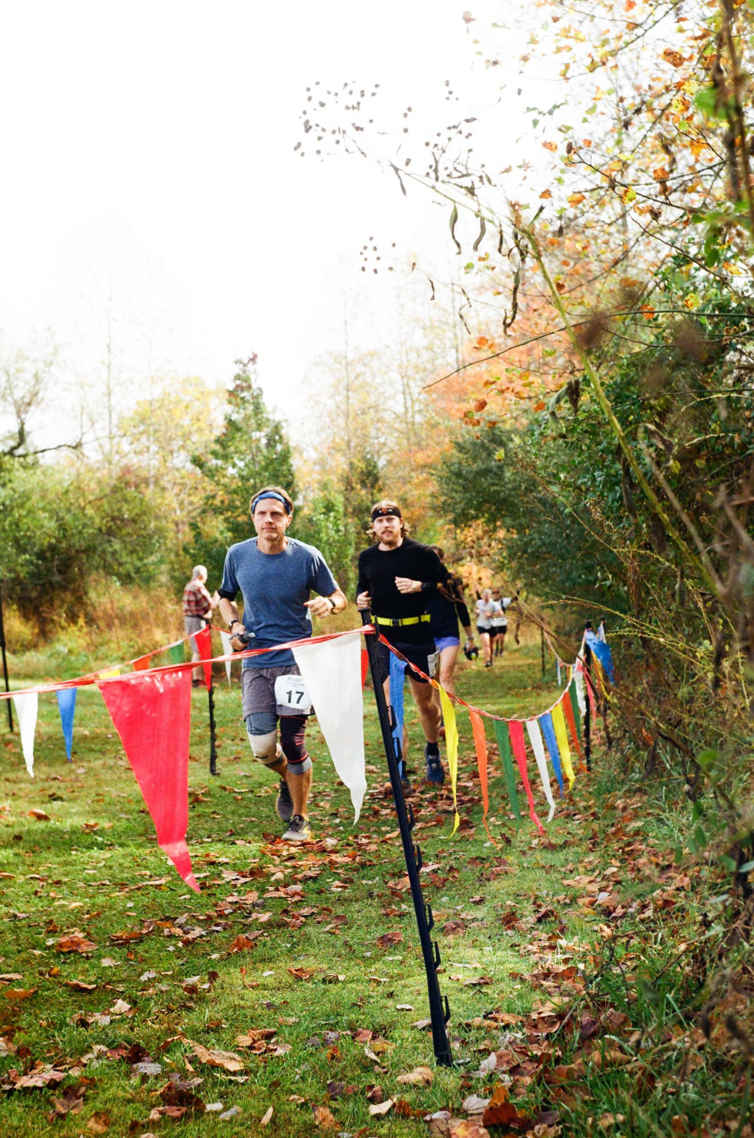 People running through an autumn obstacle course with colorful flags and fallen leaves on a grassy slope.