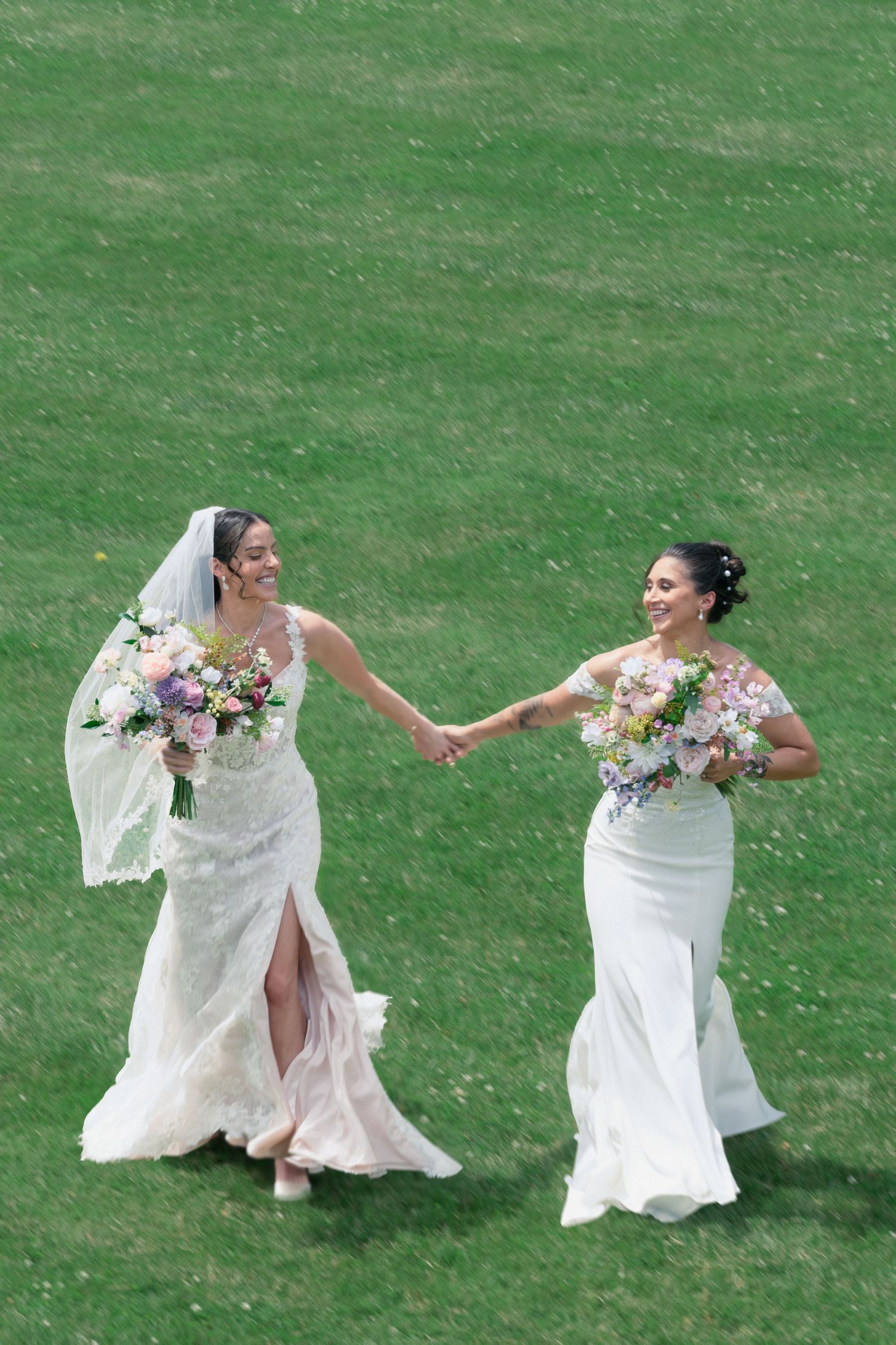 Two brides in white wedding gowns hold hands and walk across a green lawn, each carrying bridal bouquets.