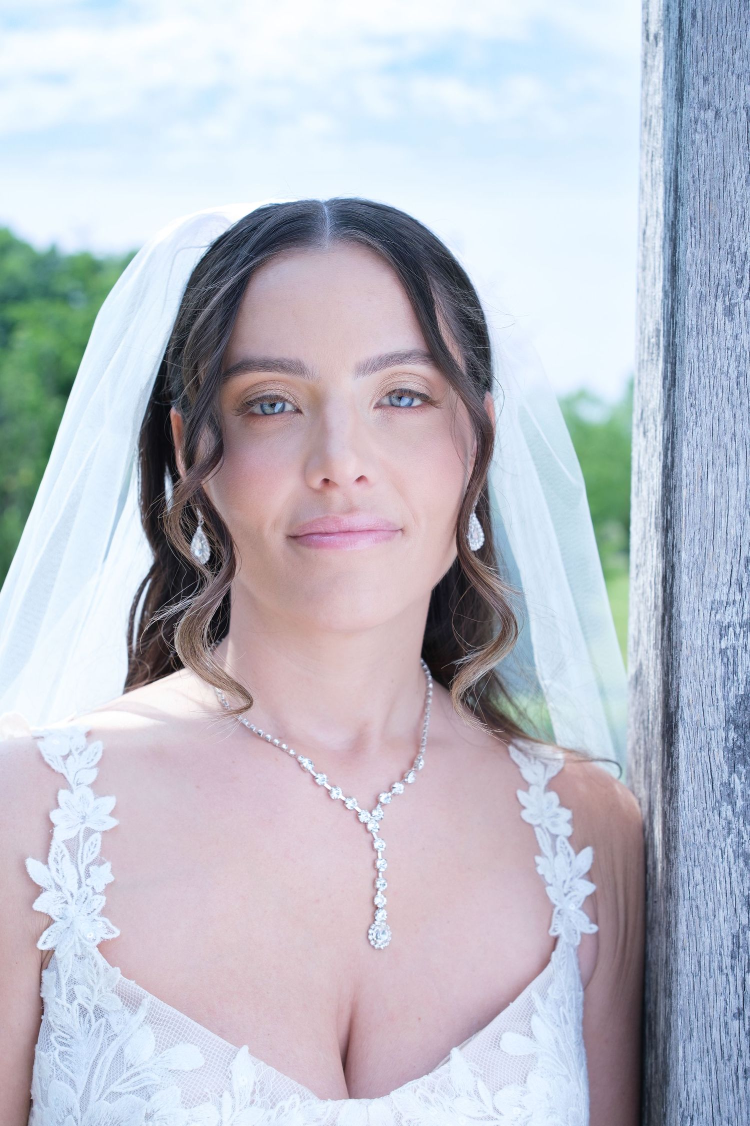 Beautiful bride wearing a lace wedding dress, diamond necklace, and white veil poses outdoors.